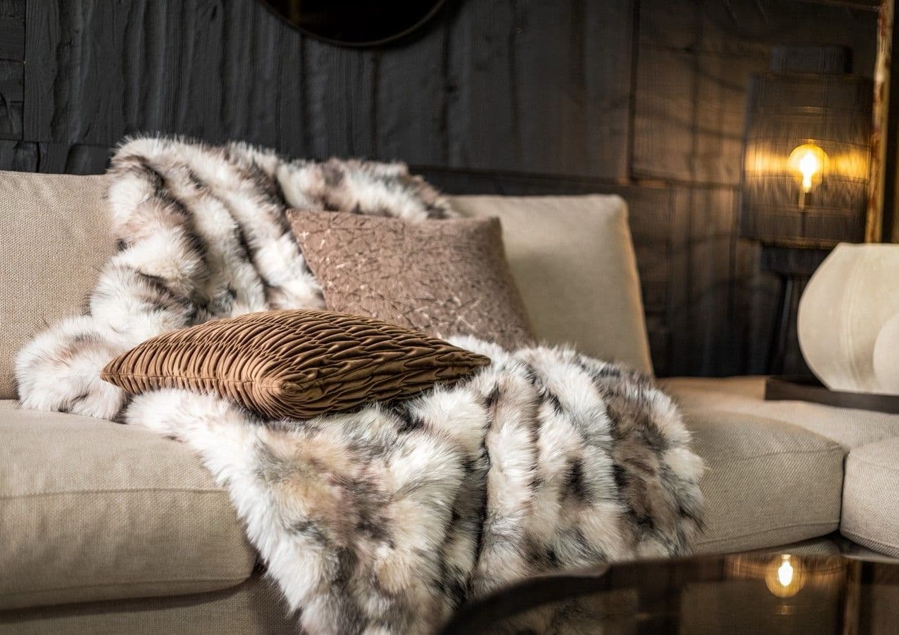 Zachte bontlook plaid in natuurtinten over een beige bank met twee sierkussens, met een warm brandende wandlamp op de achtergrond