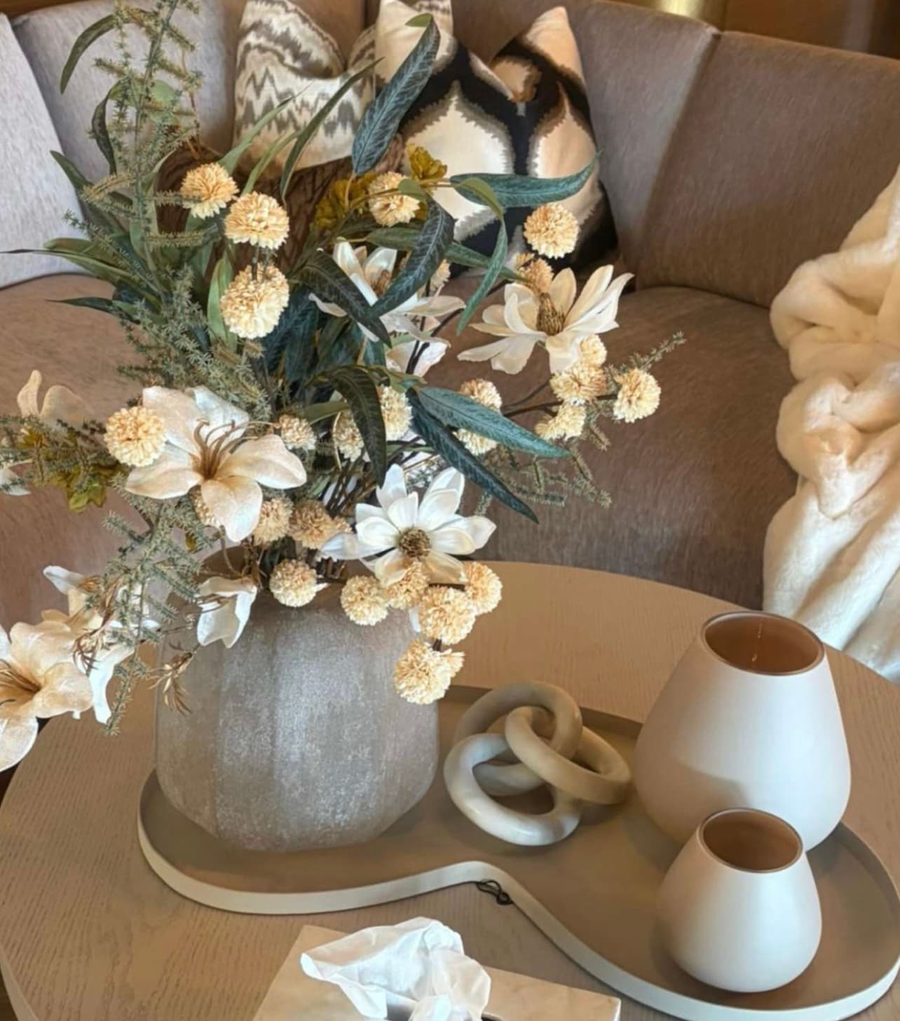 Boeket crèmekleurige zijden bloemen met groen blad in een steengrijze vaas op een houten salontafel, met een bank en een plaid op de achtergrond