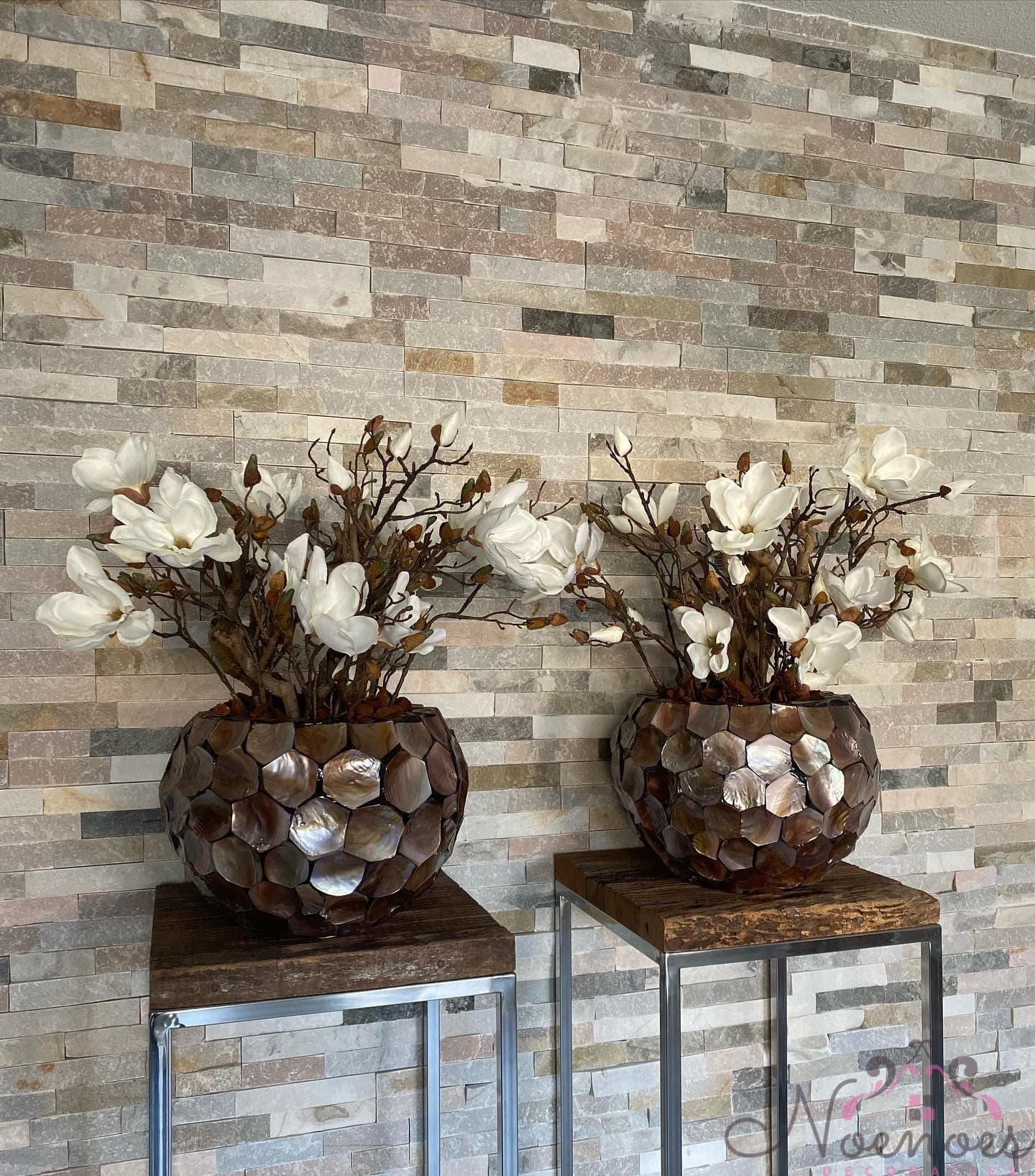 Twee ronde schelpenvazen met parelmoeren schelpjes, elk opgemaakt met witte zijden magnolia's, naast elkaar tegen een wand van natuurstenen stroken