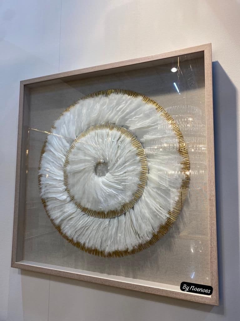 Ingelijste wanddecoratie met witte veren in een spiraalvorm met gouden randen, in een lichte houten shadowbox met glas