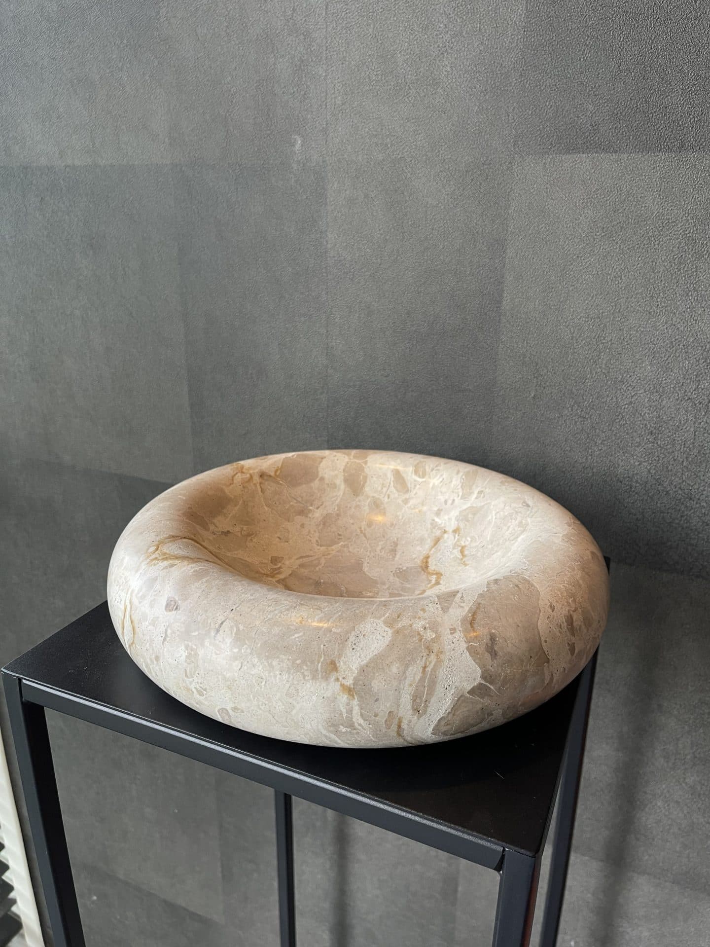 Ronde schaal van taupe en beige marmer met een dikke afgeronde rand, staand op een donker blad tegen een grijze wand