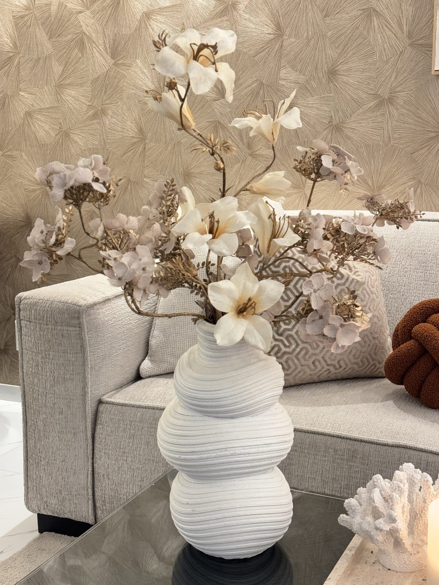 Lichte bank met beige en bruine sierkussens, een witte geribbelde vaas met crèmekleurige bloemen en een wand met bladpatroon