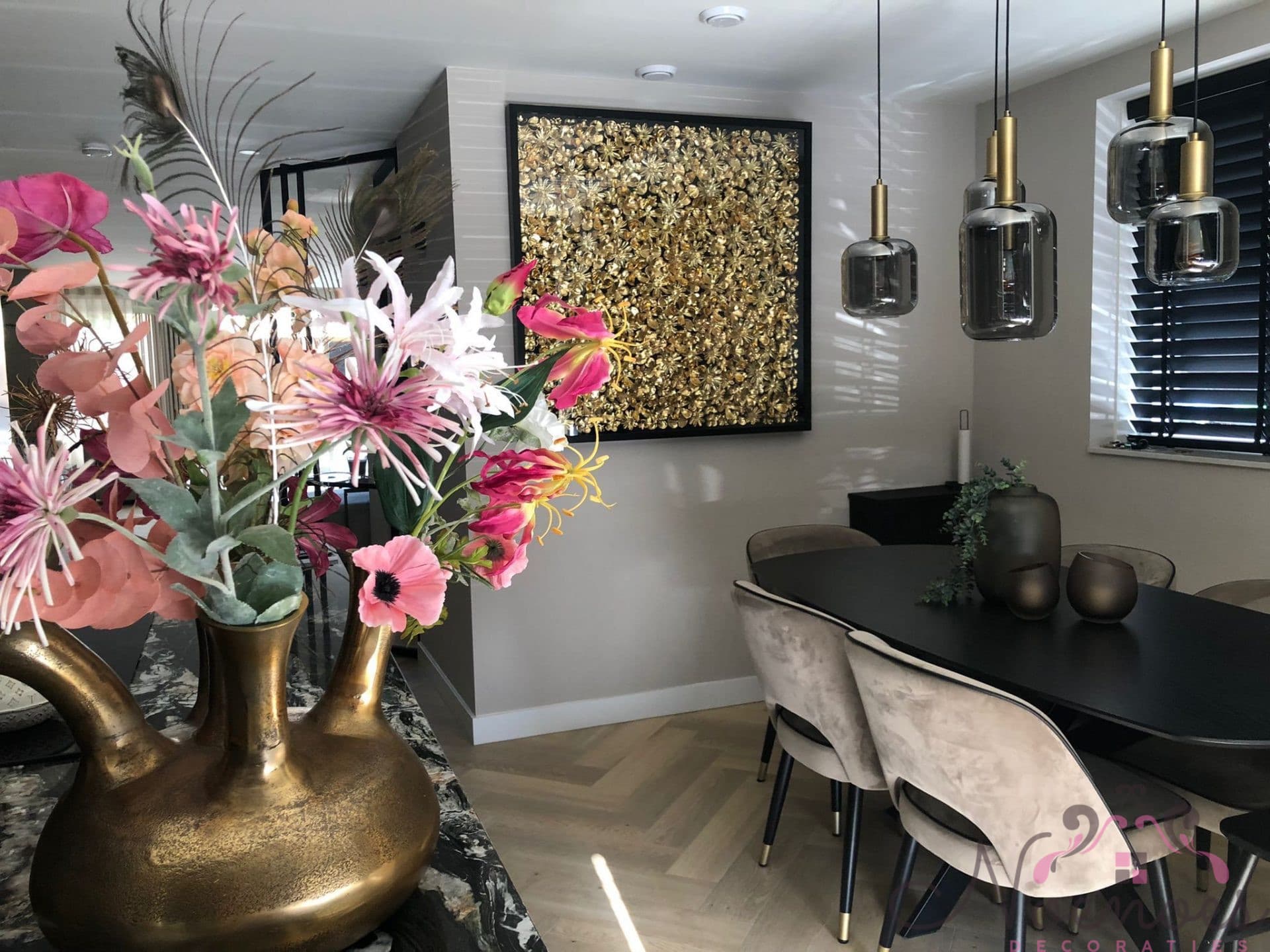 Eetkamer met een groot goudkleurig kunstwerk aan de wand, een zwarte eettafel met rookglazen hanglampen en een gouden vaas met bloemen op de voorgrond