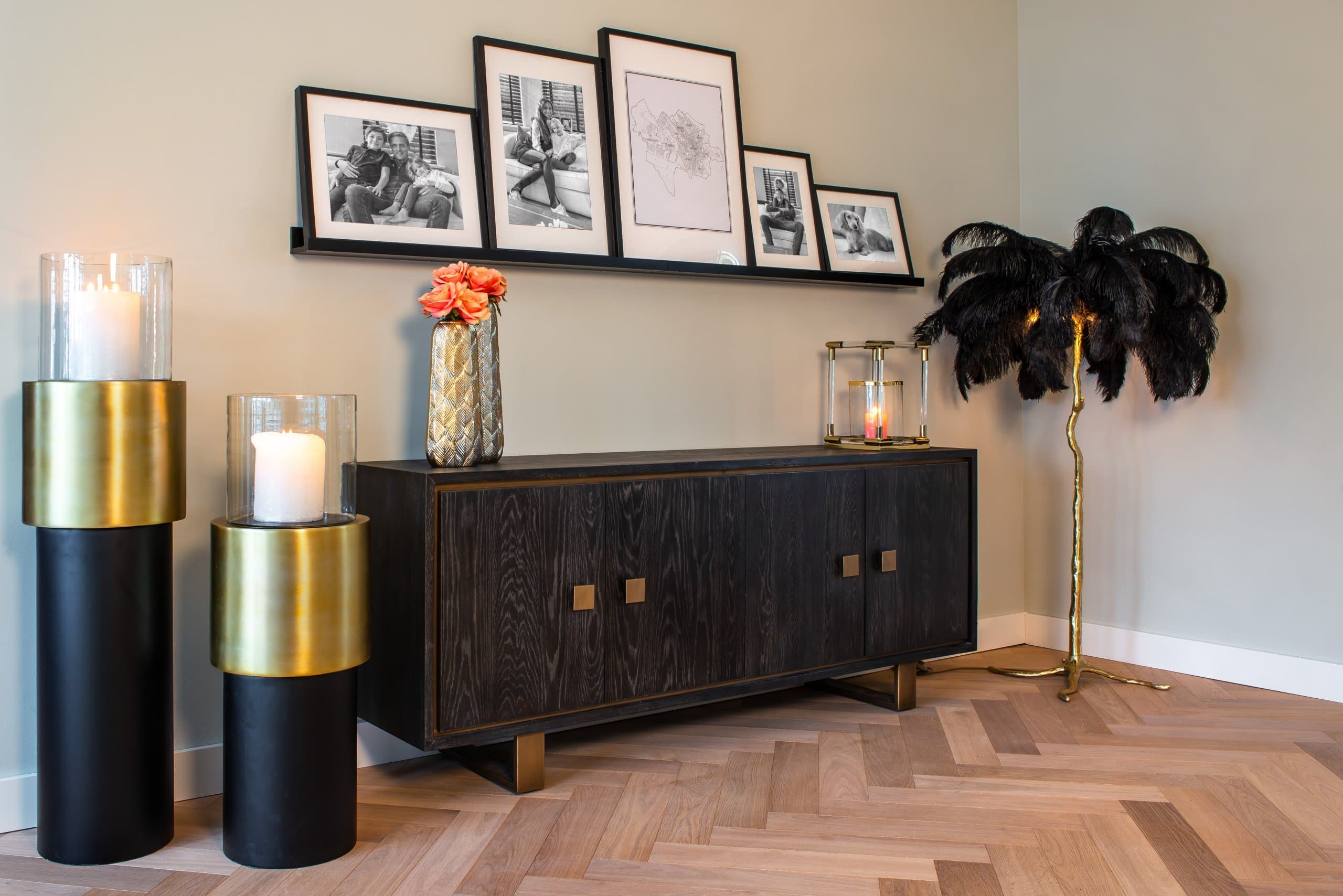 Zwart dressoir met vier deuren op een goudkleurig metalen onderstel tegen een beige wand, met visgraatparket dat eronder doorloopt en een gouden vloerlamp ernaast