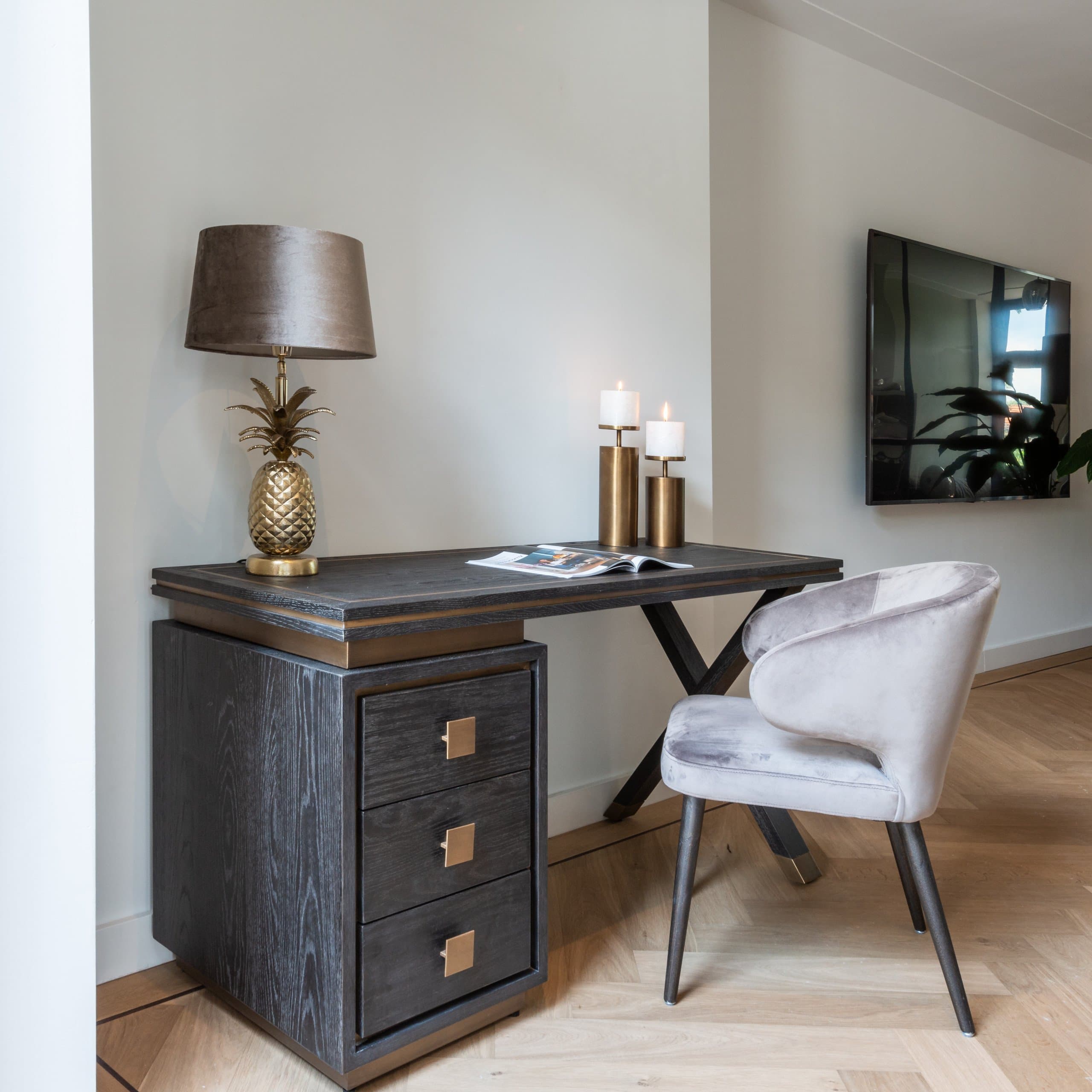 Donker houten bureau met ladeblok tegen een witte wand, met een gouden ananaslamp, twee kaarsenhouders en een lichtgrijze fauteuil ervoor