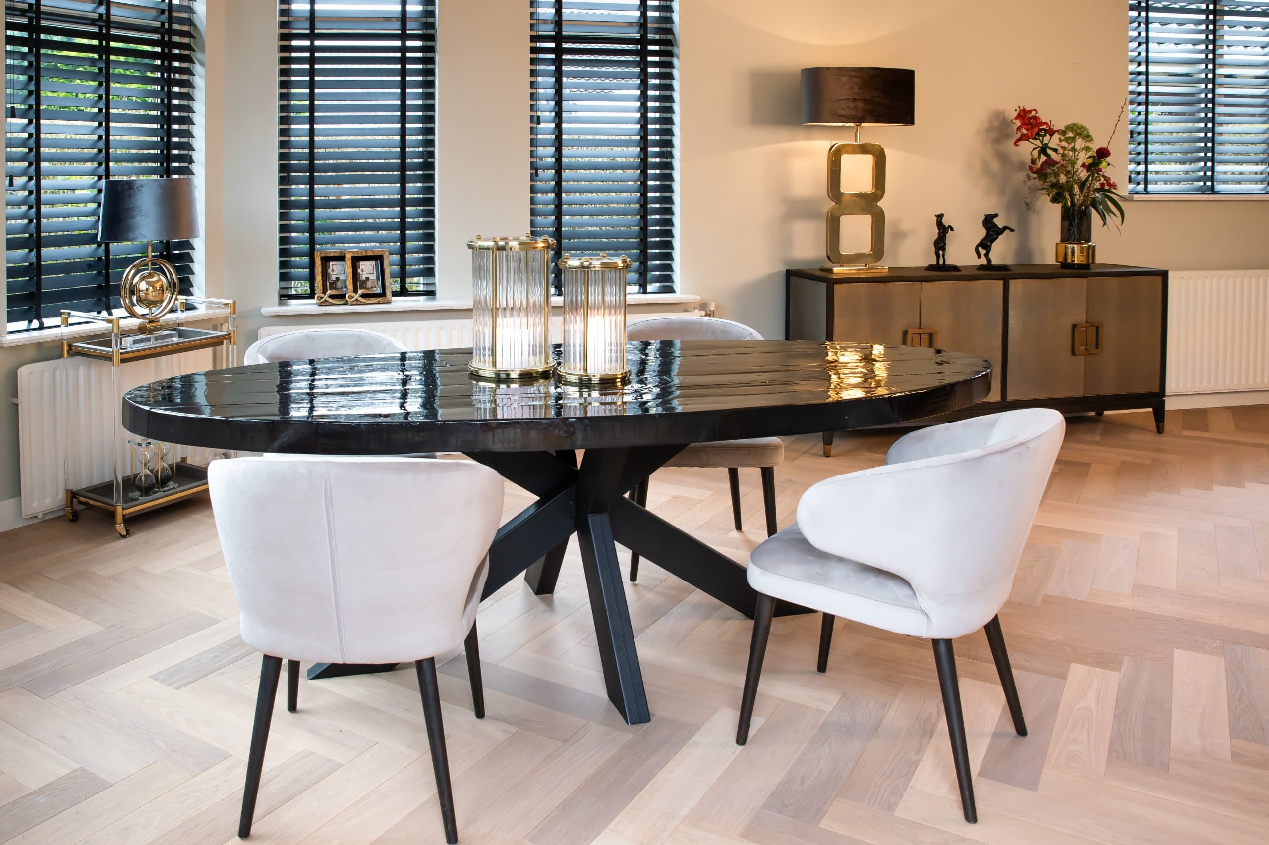 Eetkamer met een ovale zwarte tafel, lichte velvet stoelen, een donker dressoir en een fotolijst op de vensterbank tussen de jaloezieën.
