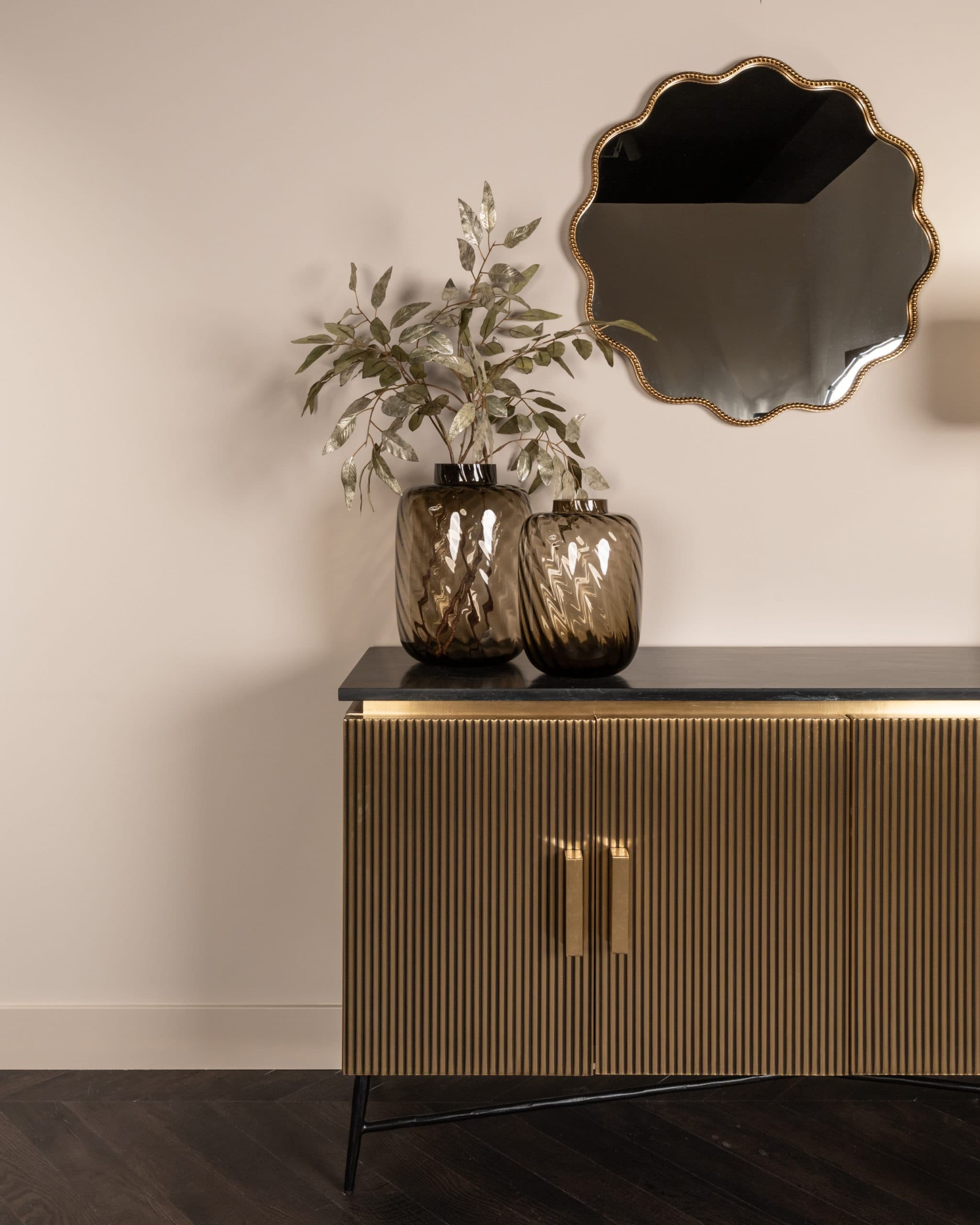 Dressoir met zwart marmeren blad en geribbeld goudkleurig front, met twee bruine glazen vazen met takken erop en een gouden spiegel erboven