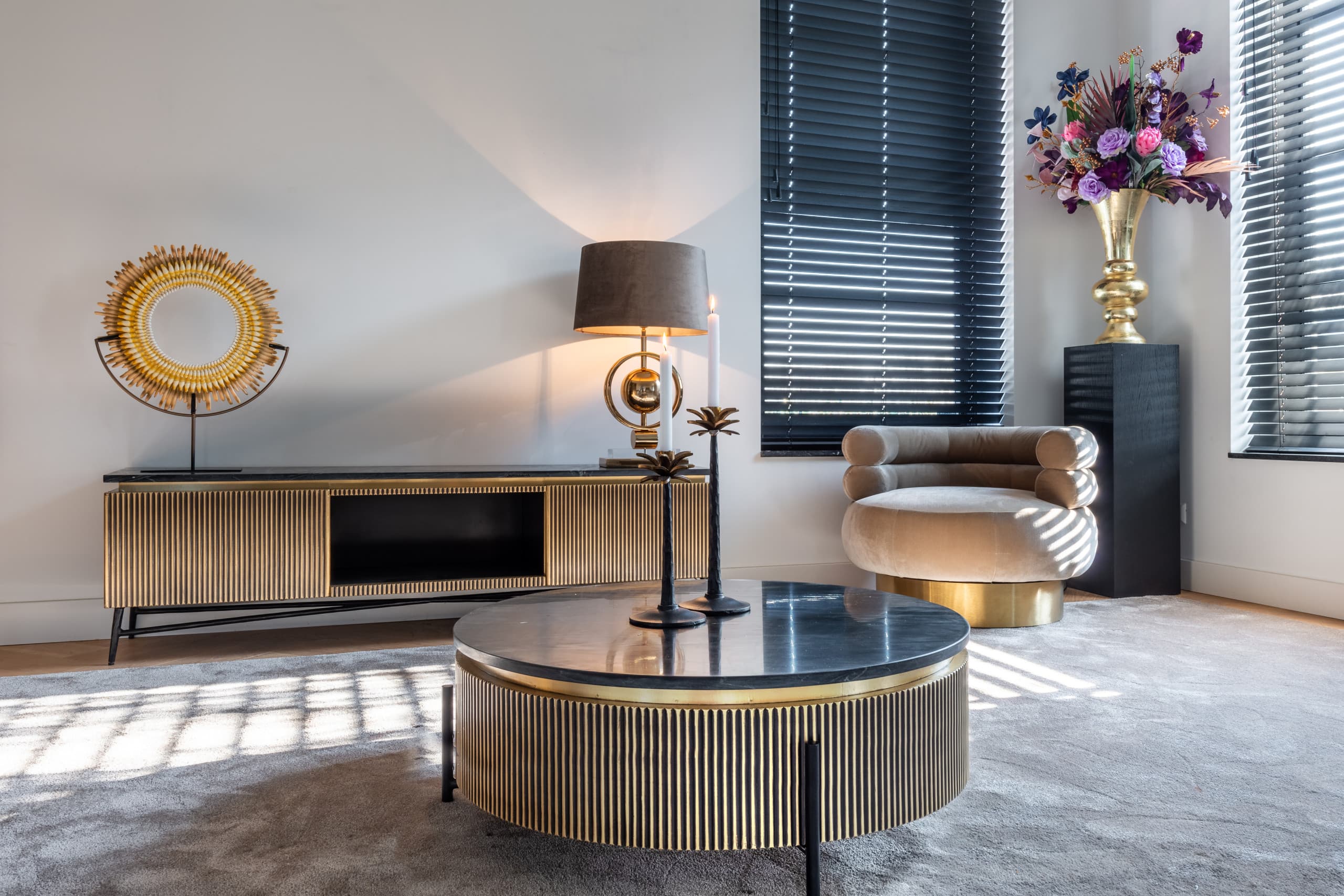 Woonkamer met een goudkleurig tv-meubel waarop een tafellamp en een rond zonnesculptuur staan, met een ronde salontafel en een fauteuil bij het raam.
