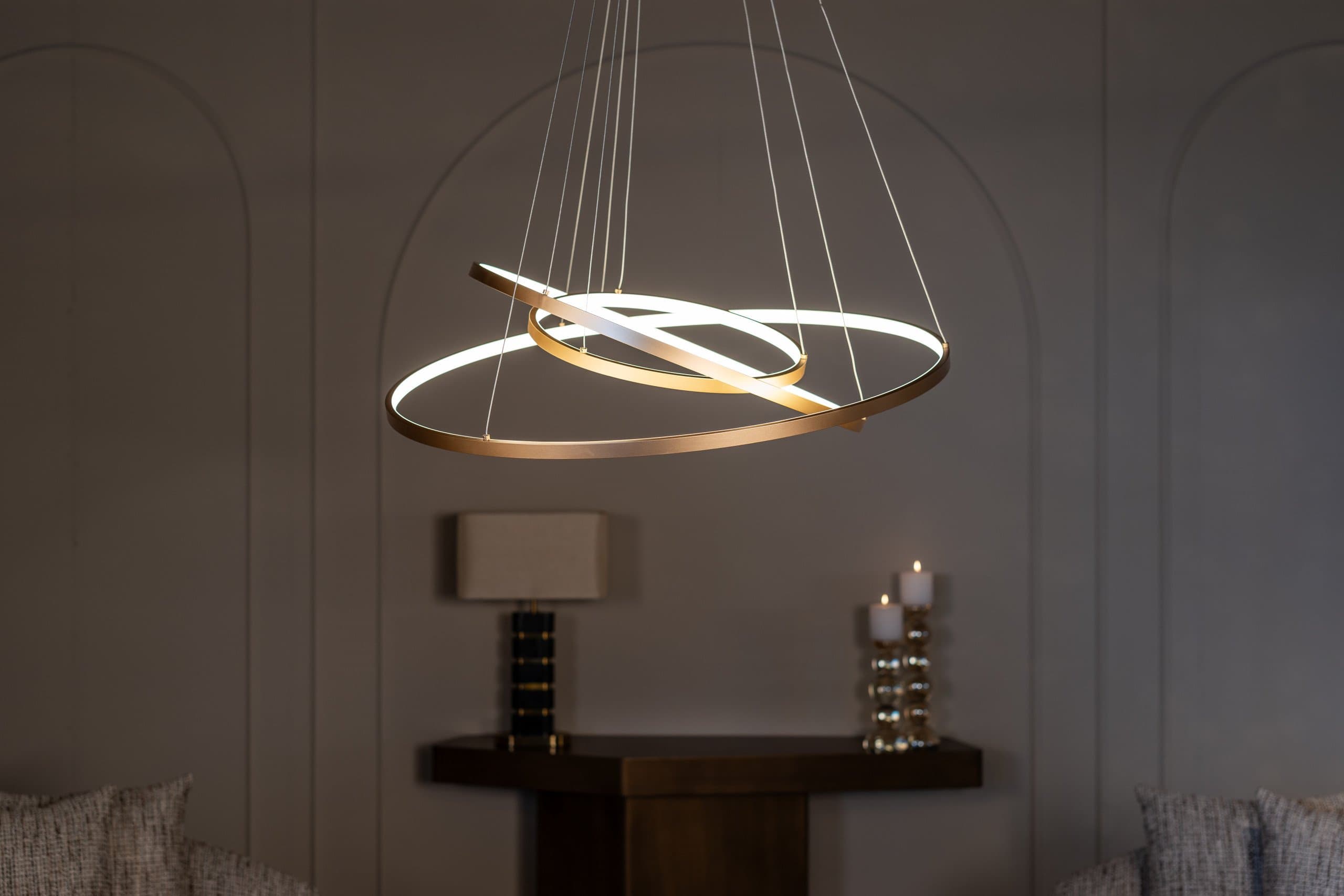 Goudkleurige hanglamp met twee kruisende lichtringen boven een donker dressoir met tafellamp en kaarsen