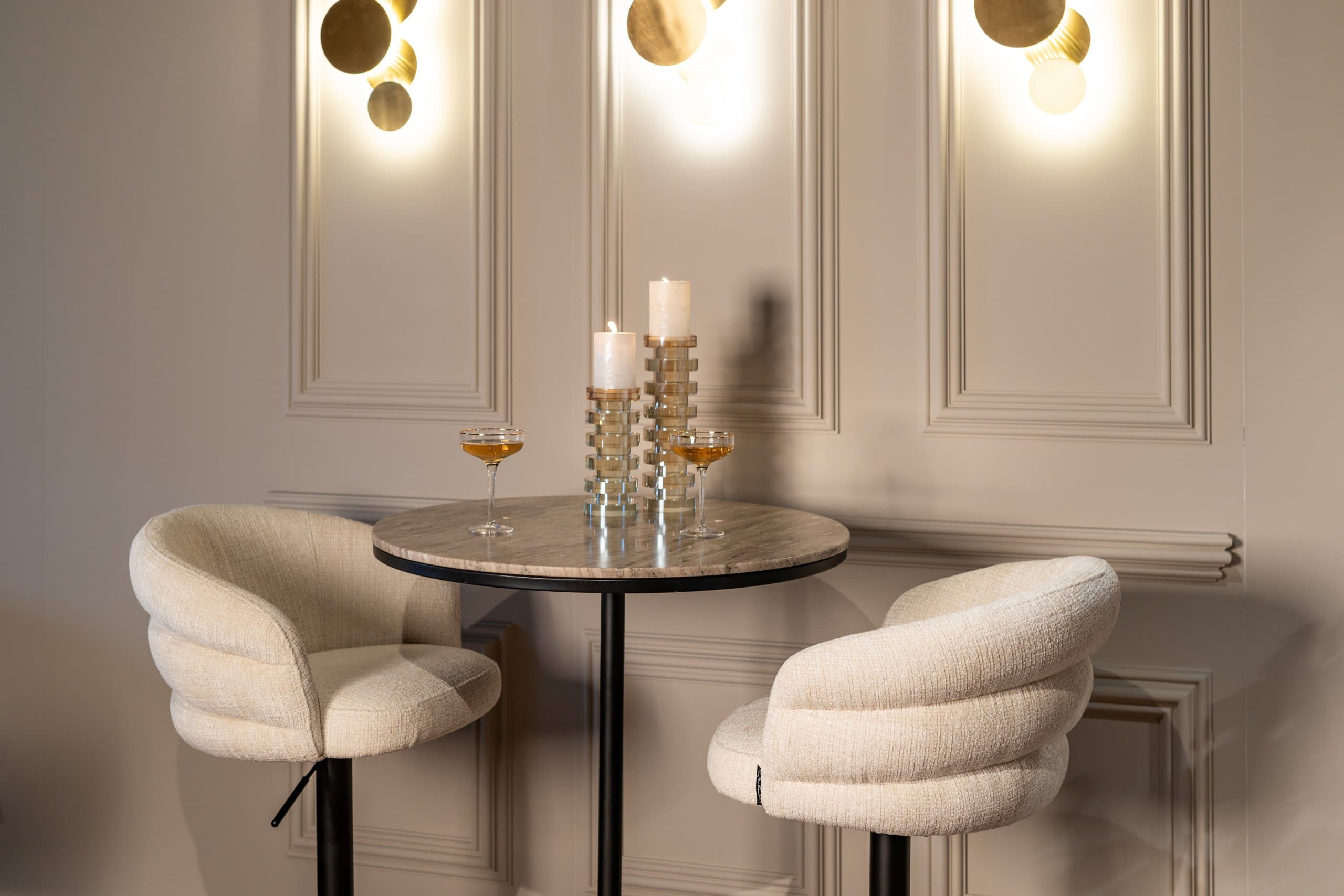 Wand met gebroken witte panelen en lijstwerk, drie ronde wandlampen in goud, een ronde marmeren tafel met kaarsen en twee crèmekleurige bouclé stoelen