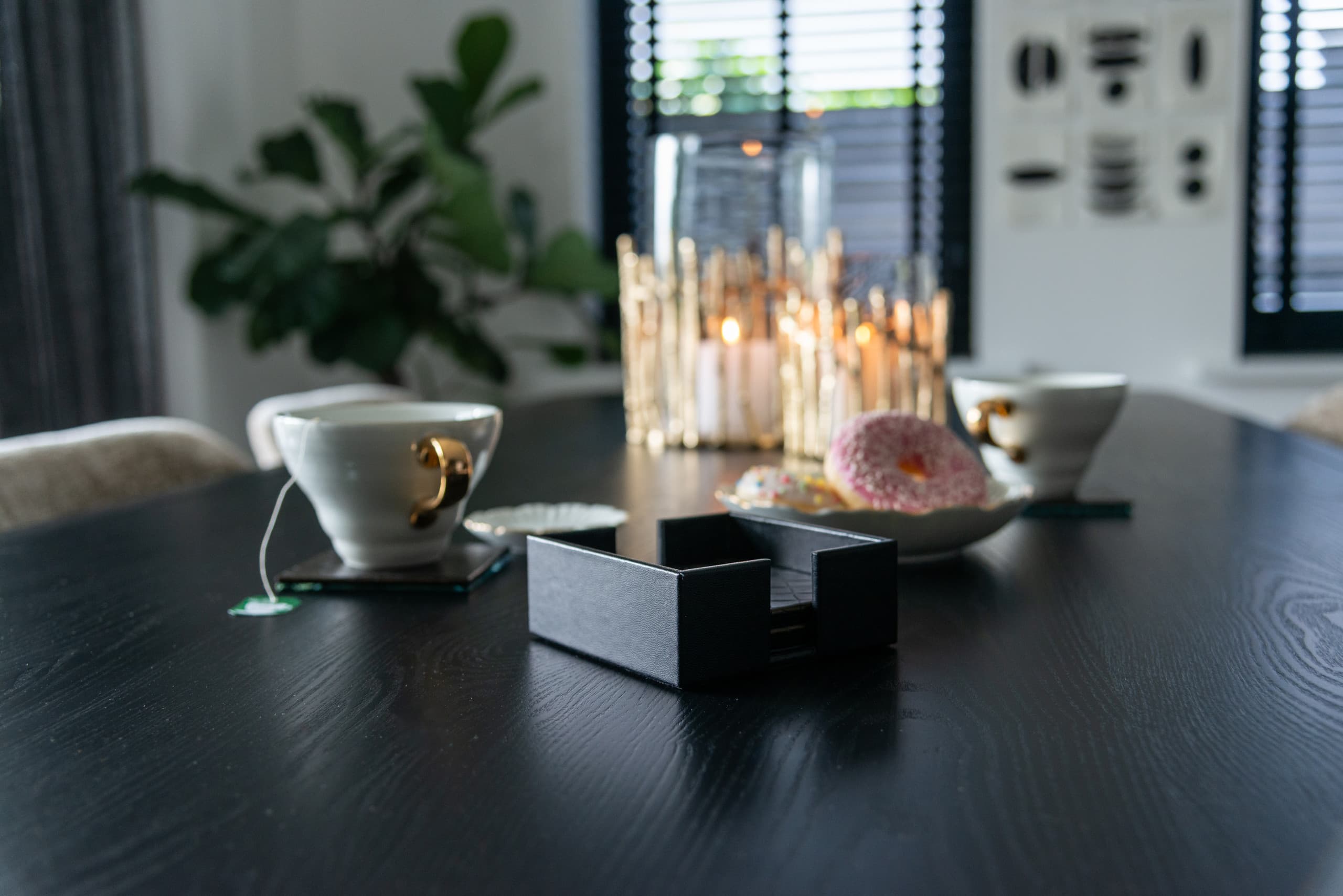 Twee glazen windlichten met brandende kaarsen op een donkere houten eettafel, met twee kopjes en een schaaltje op de voorgrond