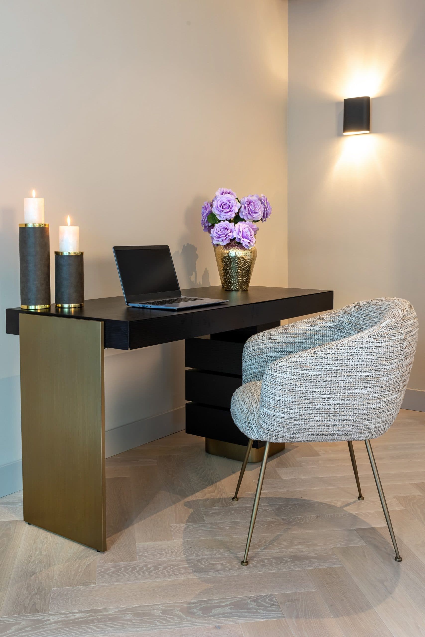 Donker bureau met laden en gouden zijpaneel, met opengeklapte laptop, een gouden vaas met paarse rozen en een lichte gestoffeerde stoel ervoor