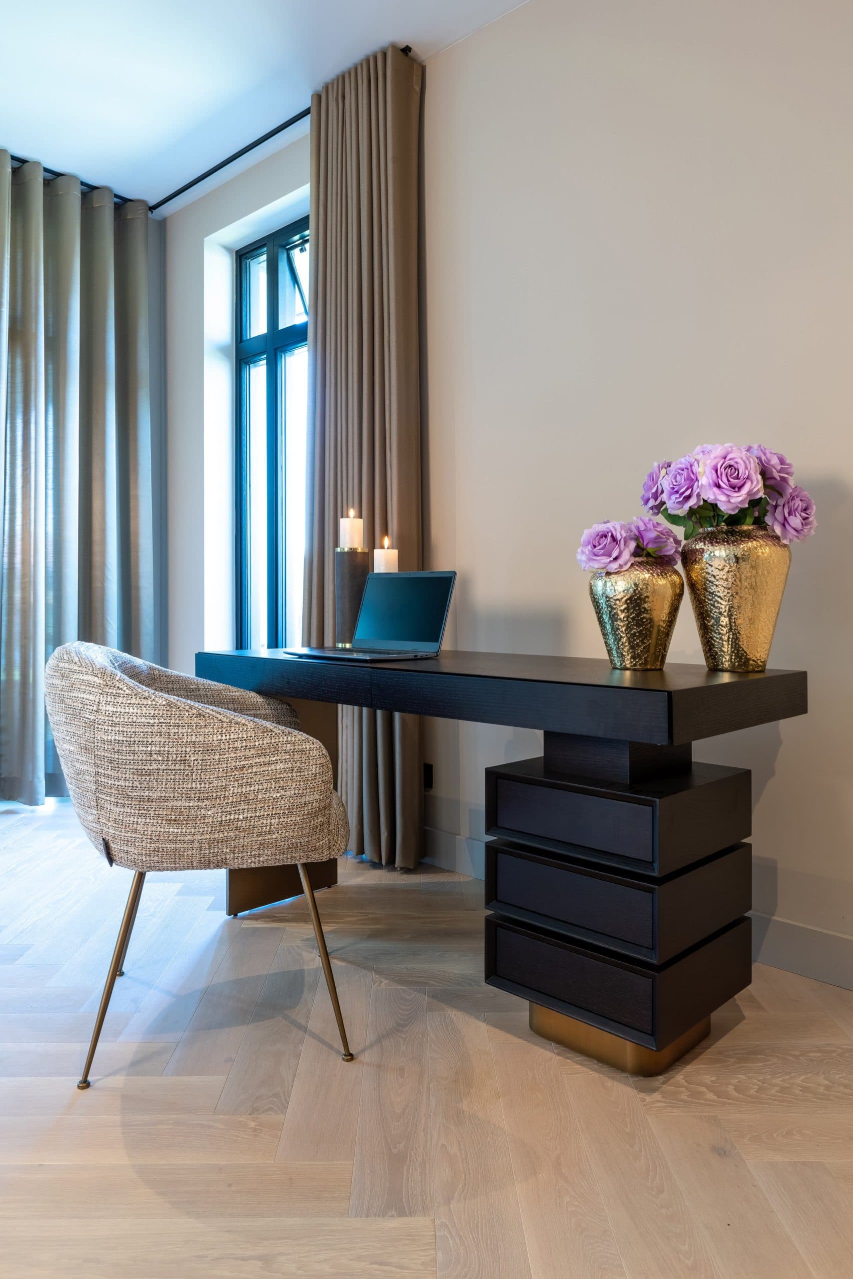 Donker houten bureau met een laptop, twee gouden vazen met paarse rozen, brandende kaarsen en een gestoffeerde stoel voor een raam met gordijnen.