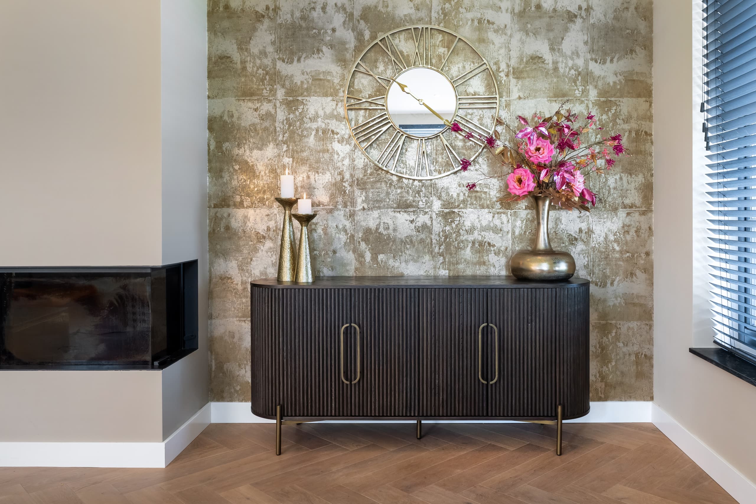 Breed dressoir van donker geribbeld eiken op gouden poten tegen een goudkleurige wand, met twee kandelaars, een messing vaas met roze bloemen en een ronde klok erboven