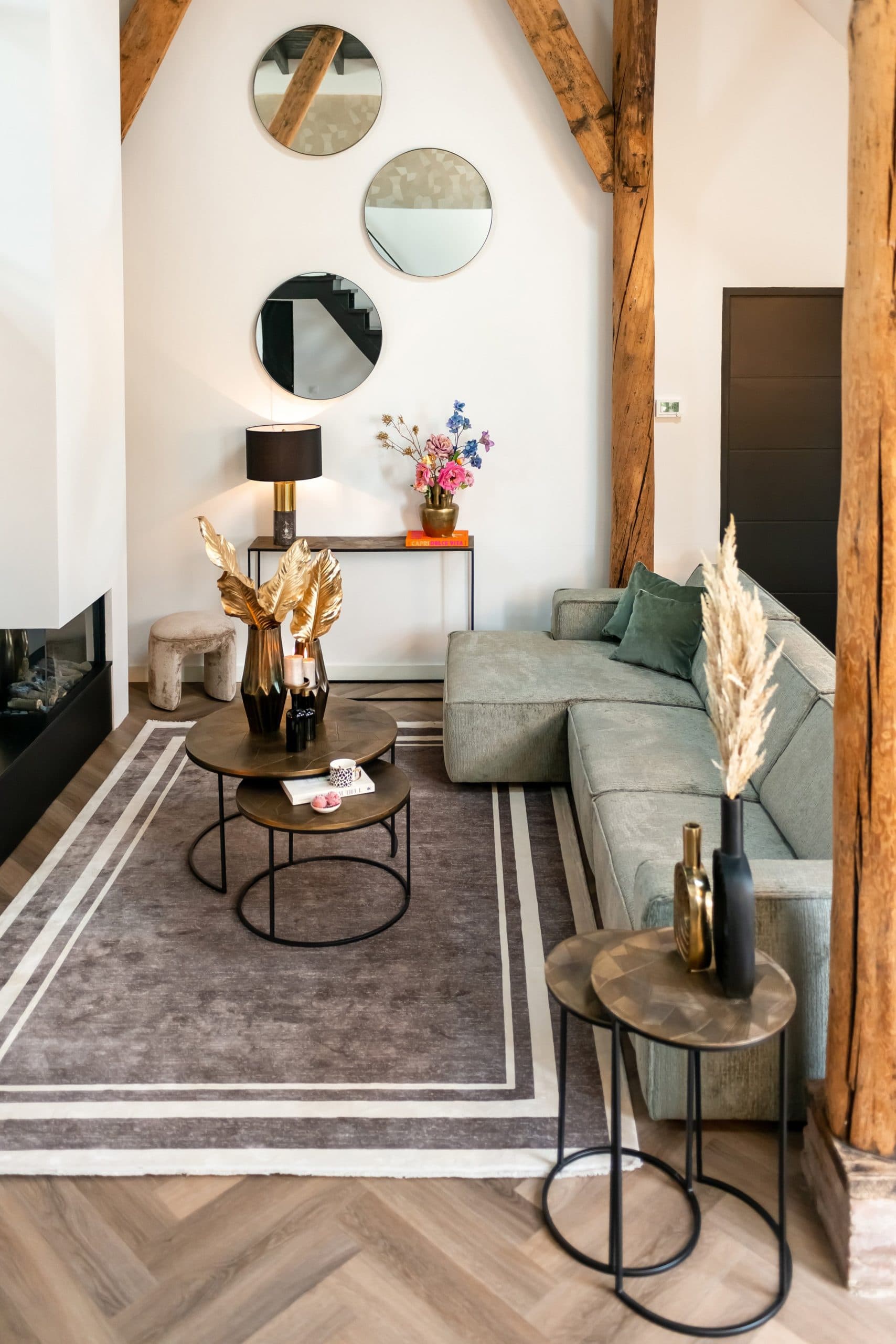 Twee ronde geneste bijzettafels met bronskleurig blad op een grijs vloerkleed voor een groene loungebank, met een smalle console en zwarte tafellamp tegen de witte wand