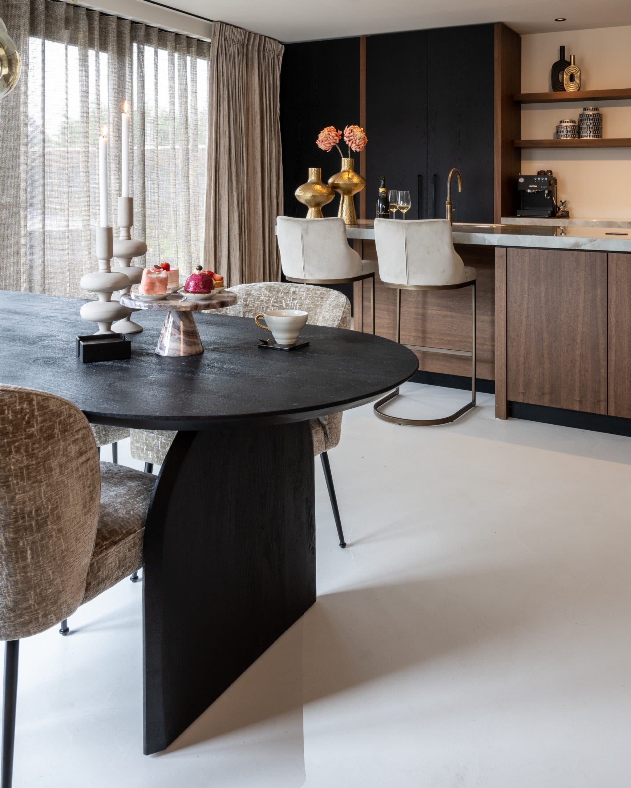 Ovale zwarte eettafel op de voorgrond met daarachter een kookeiland van walnoot met marmeren blad en twee lichte barstoelen