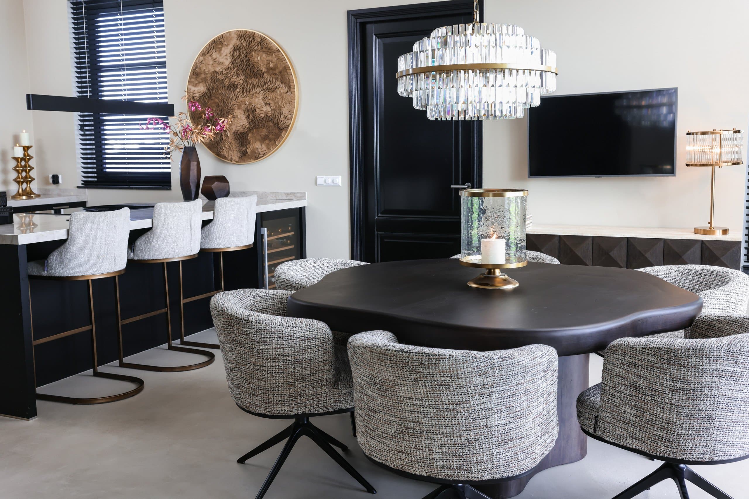 Bar met een licht stenen blad en drie gestoffeerde barkrukken op bronzen poten, met op de voorgrond een eettafel met gemêleerde stoelen