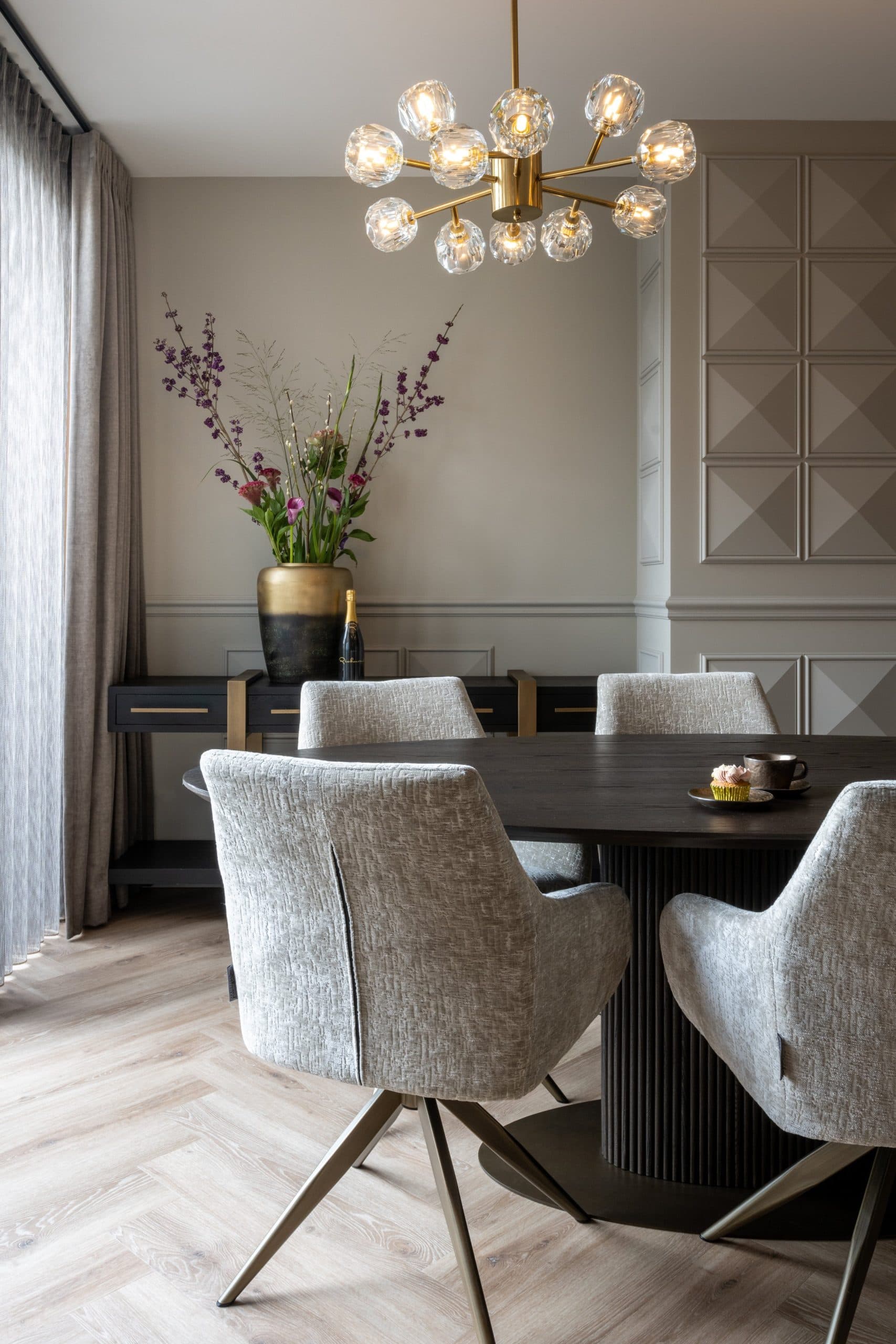 Eetkamer met een donker dressoir op slanke poten tegen een lichte lambriseringswand, een gouden vaas met bloemen erop en lichte gestoffeerde eetkamerstoelen op de voorgrond