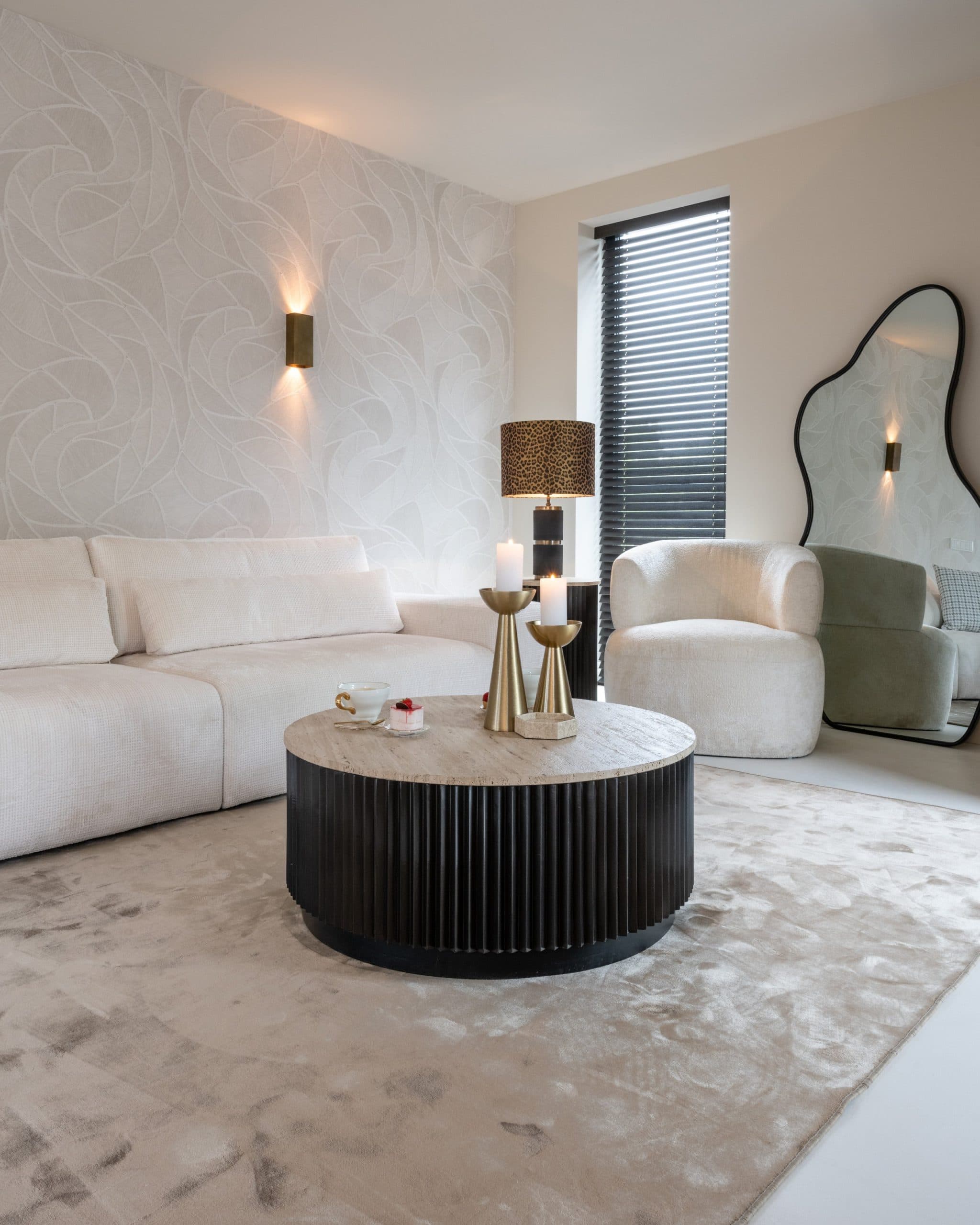 Woonkamer met een brede witte bank, een ronde salontafel met geribbelde voet en rechts een grote spiegel met golvende zwarte lijst