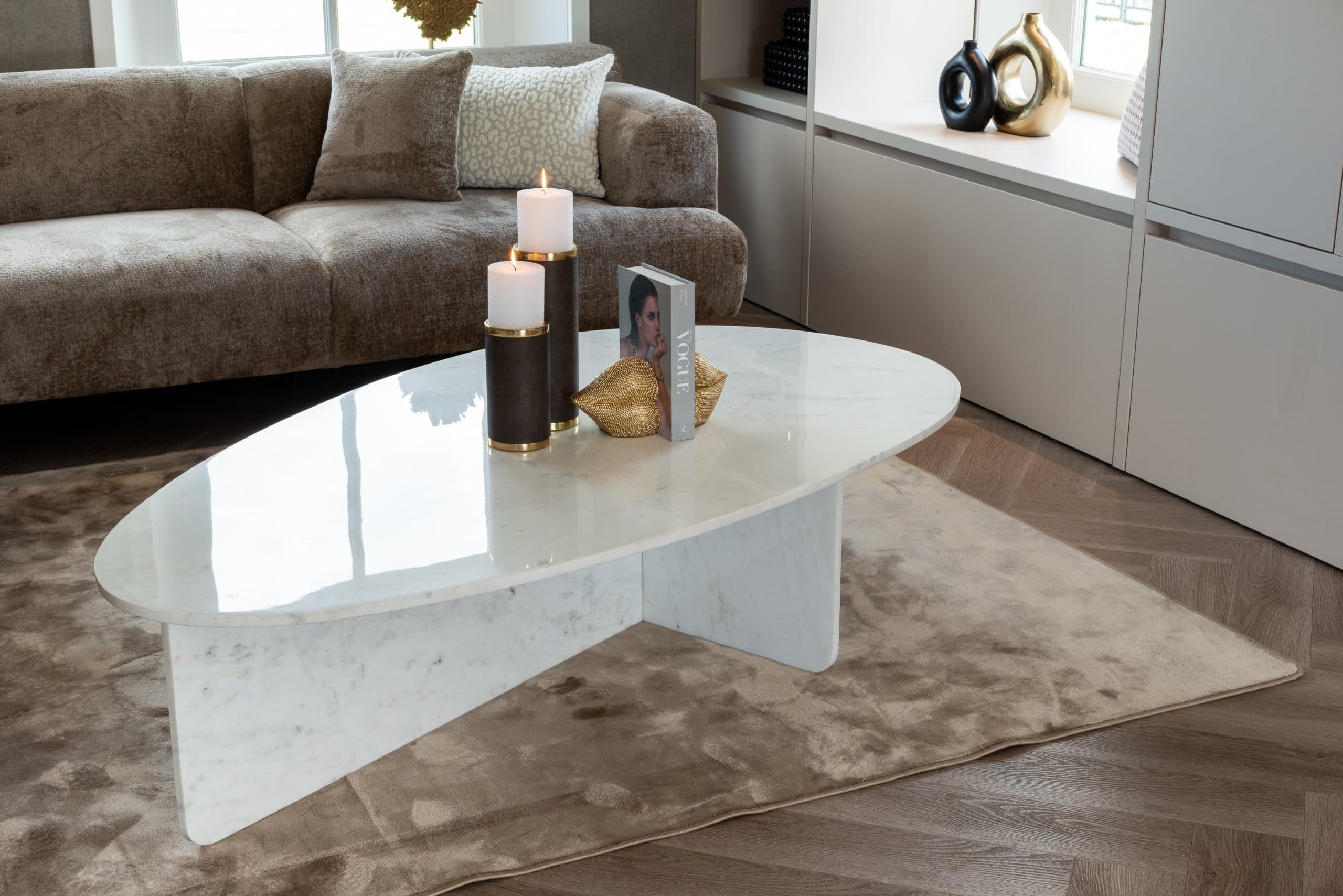 Ovale witte marmeren salontafel met twee kaarsen in houders met goudkleurige ringen en een gouden schelpvormig object naast een boek