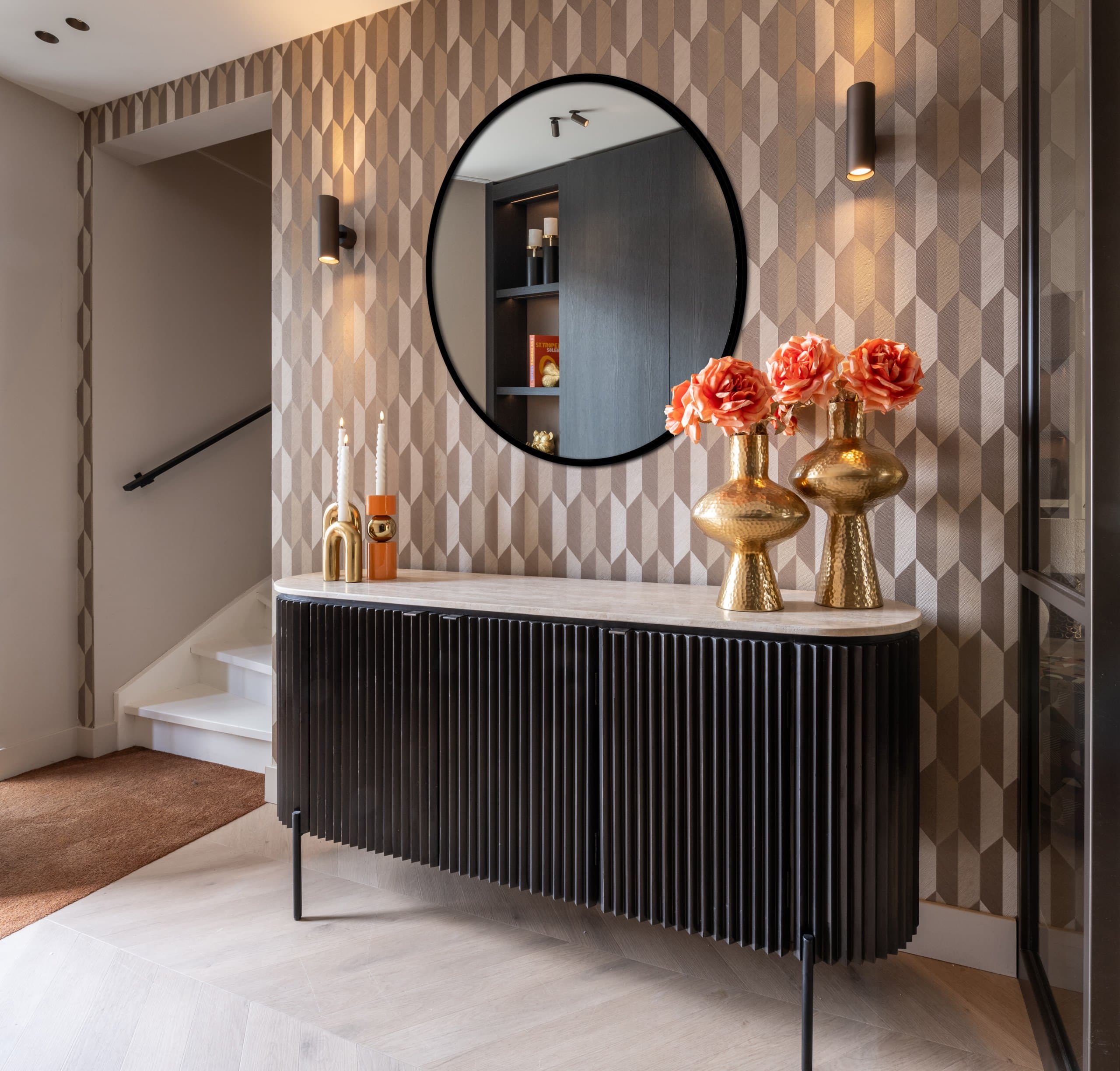 Zwart geribbeld dressoir met lichte stenen bovenblad tegen een gedessineerde wand, met twee gouden vazen met oranje rozen, kaarsen en een ronde spiegel erboven