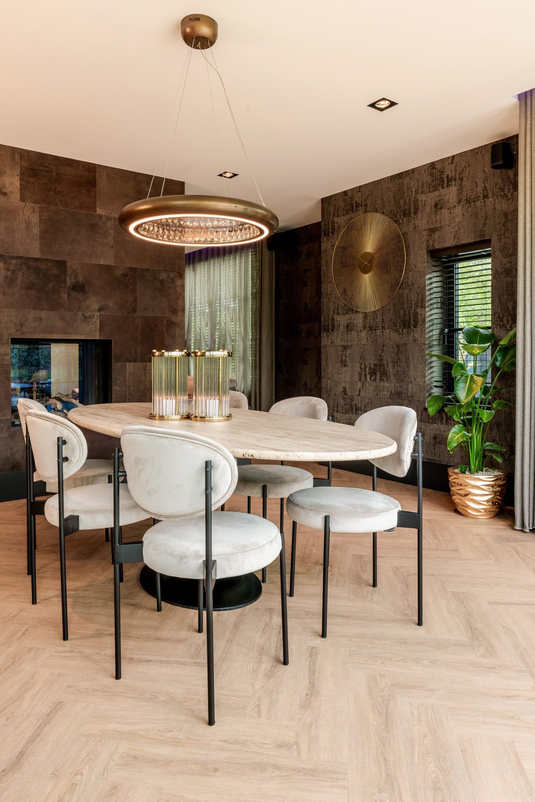 Eetkamer met ovale travertijnen tafel, lichte stoelen en een raam met zwarte jaloezieën en een gordijn ernaast