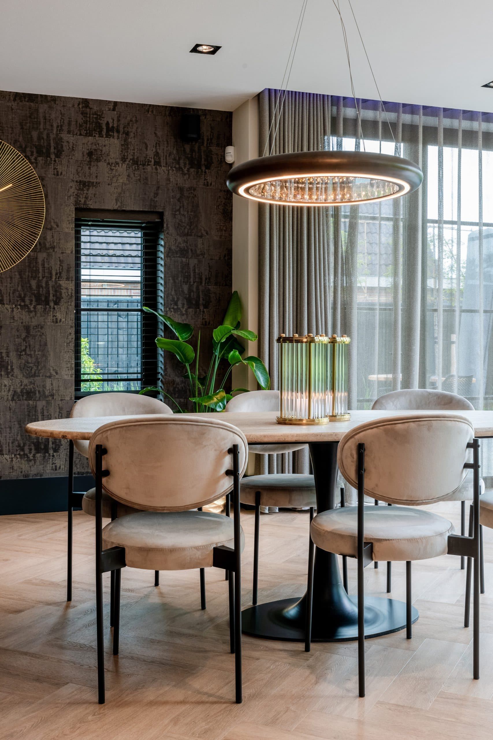 Eettafel met lichte stoelen voor een groot raam met beige gordijnen, jaloezieën en een grote groene plant
