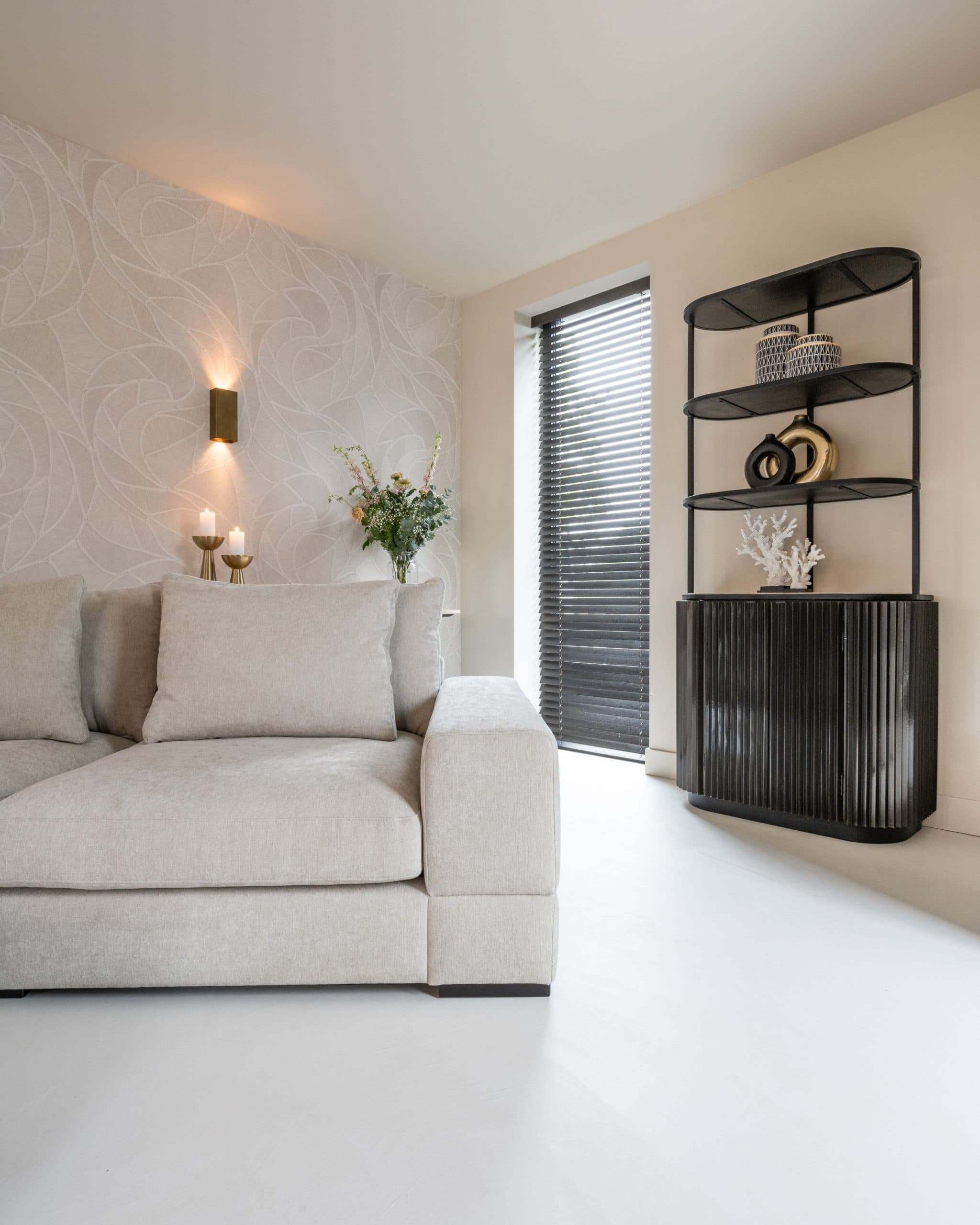 Hoge zwarte kast met geribbelde deuren en open planken erboven, met een wit koraalobject en twee ronde vazen, naast een lichte bank bij een raam met jaloezieën