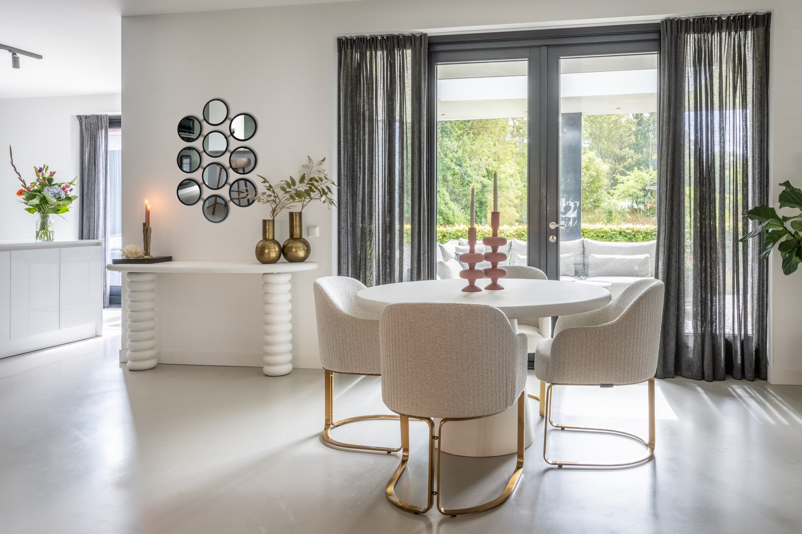 Lichte langgerekte woonkamer met een witte consoletafel langs de wand, een spiegelcompositie erboven, een ronde eettafel met vier beige stoelen en tuindeuren met grijze gordijnen