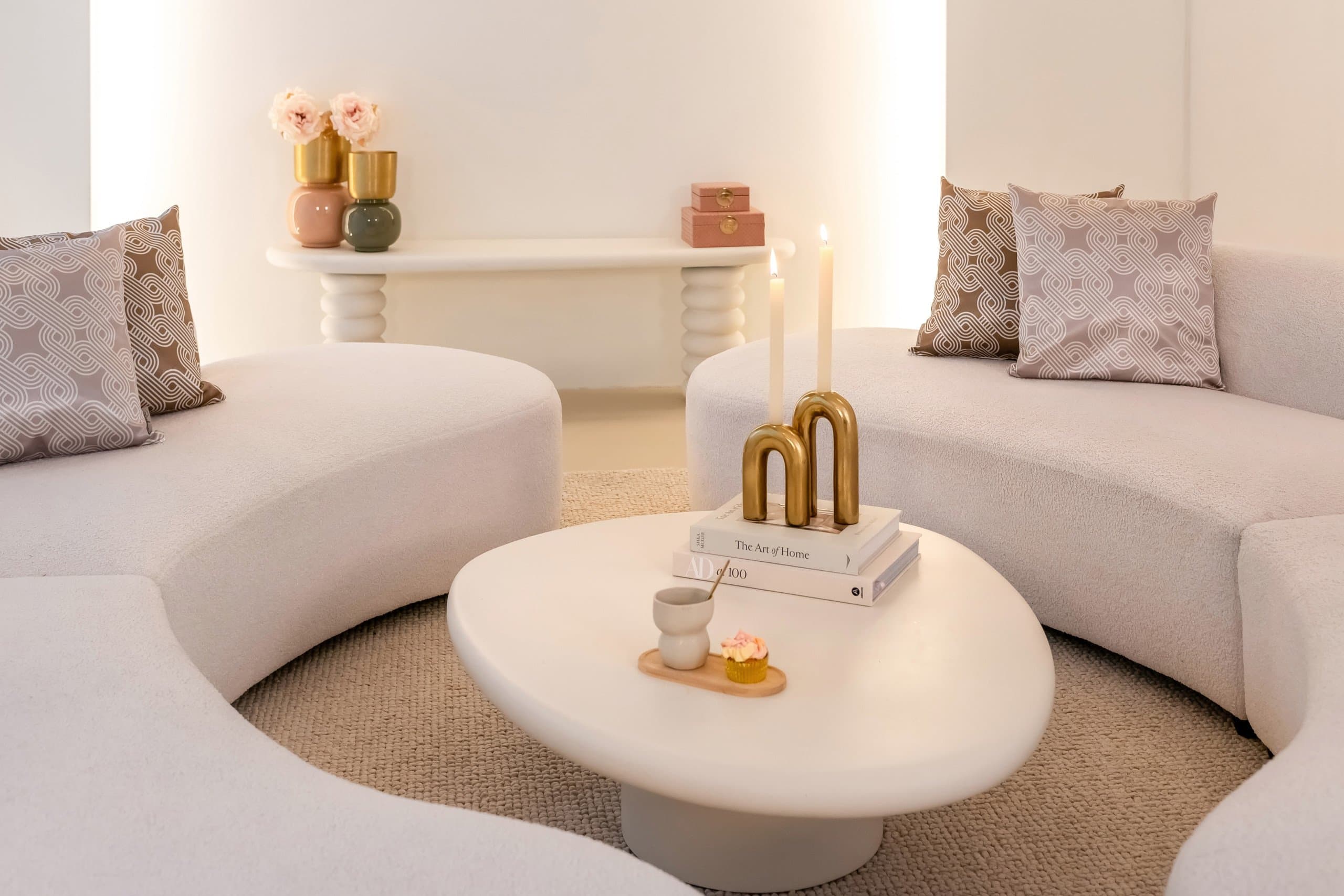 Lichte woonkamer met twee gebogen crèmekleurige banken, een organisch gevormde witte salontafel en gouden kandelaars op een stapel boeken