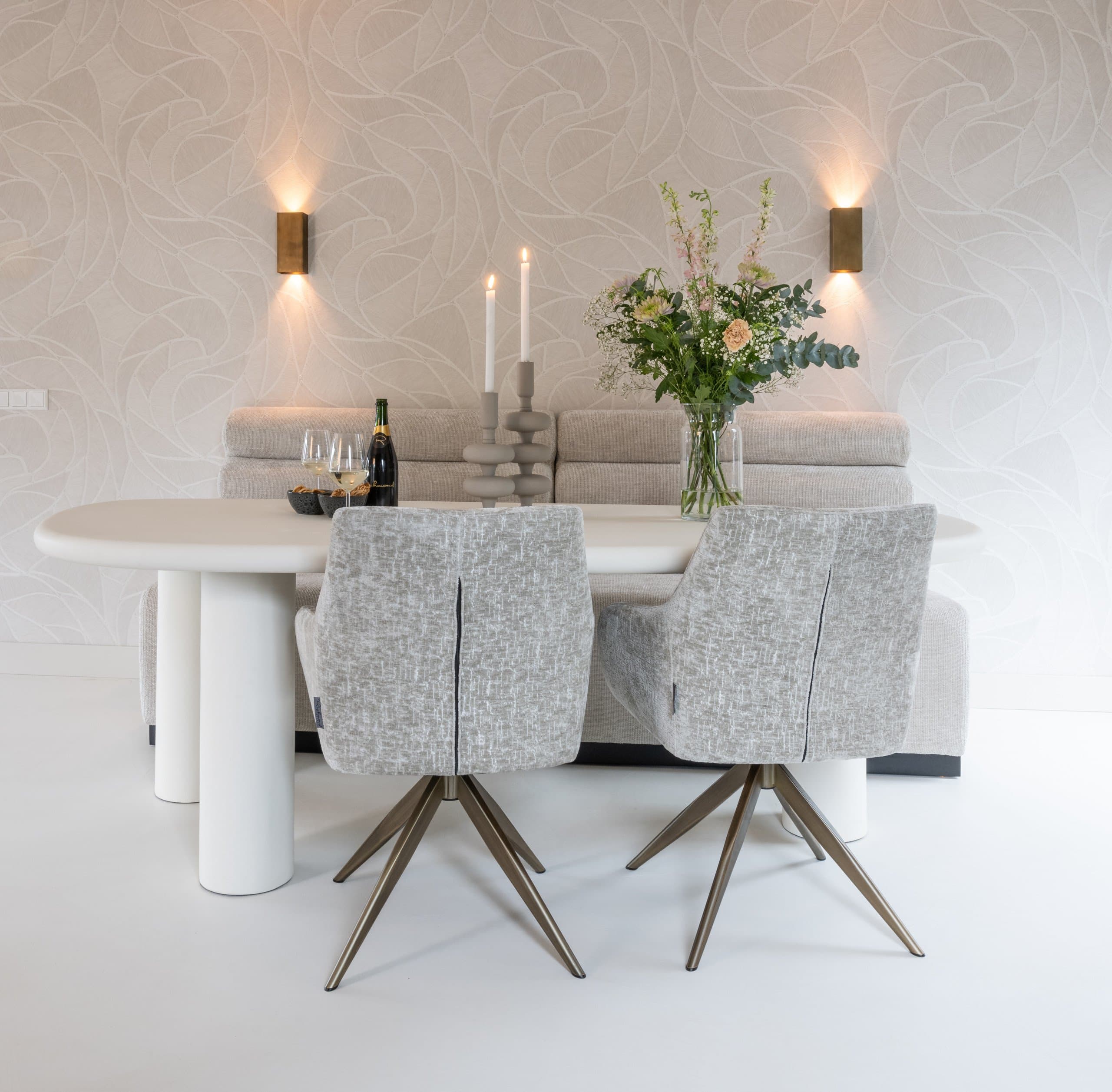 Lichte ovale eettafel met twee grijze draaistoelen, kaarsen en een vaas bloemen, tegen een wand met bladmotief
