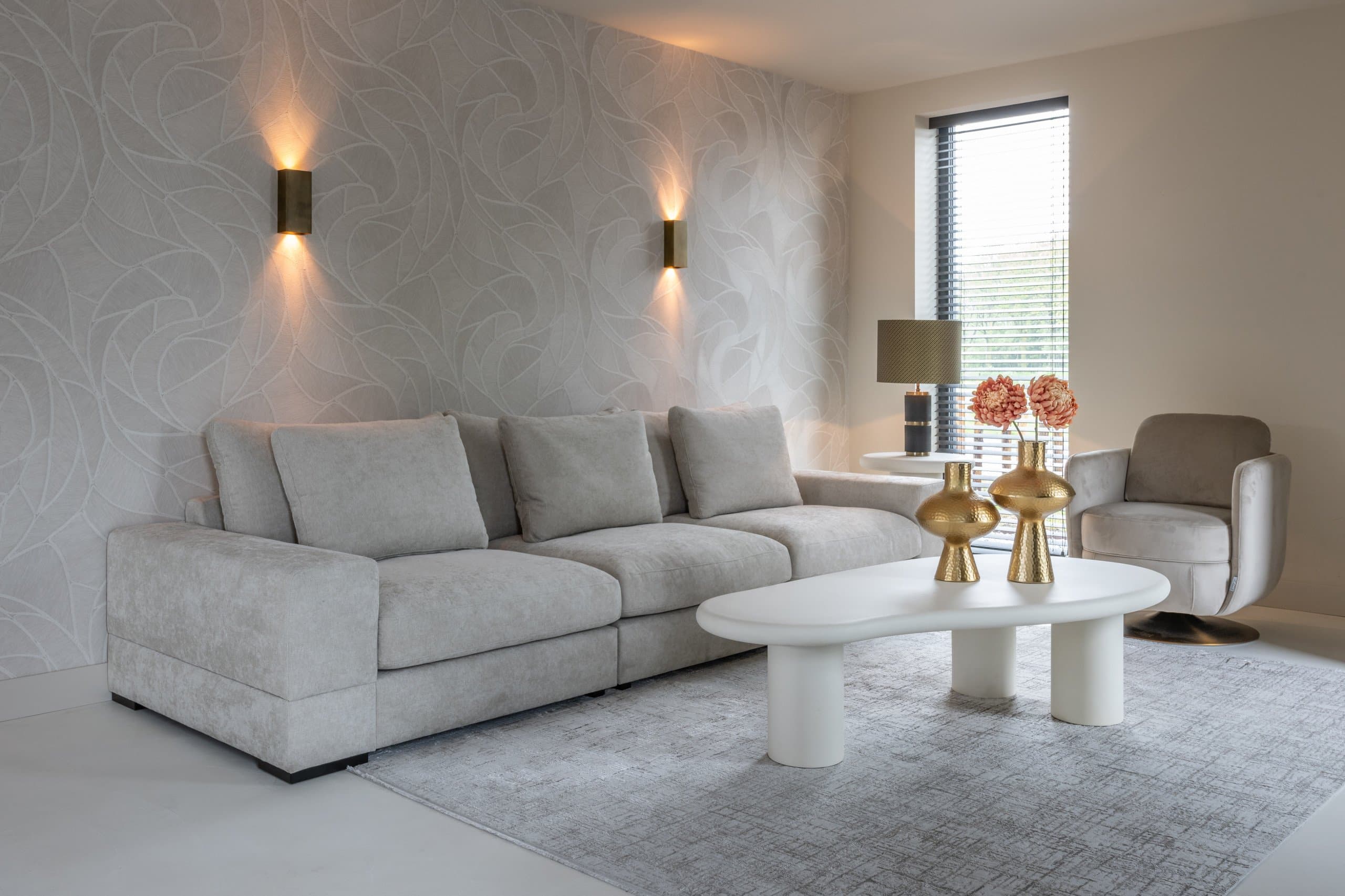 Lichte woonkamer met een crèmekleurige hoekbank, twee warme wandlampen, een witte organisch gevormde salontafel met twee gouden vazen en een fauteuil bij het raam