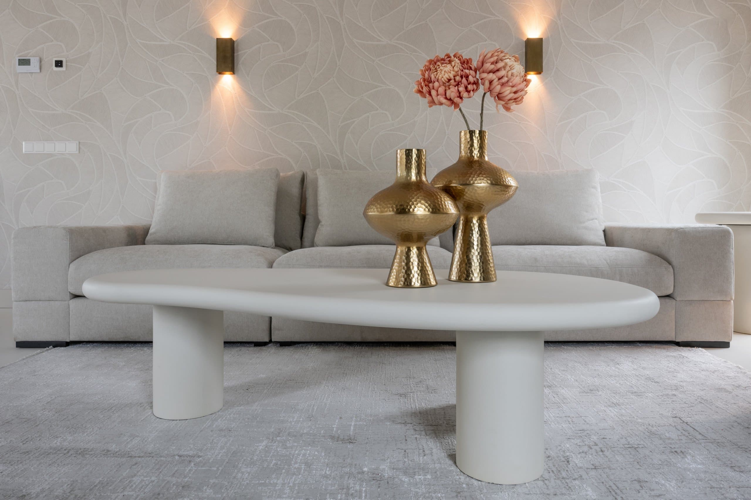 Lichtgrijze bank met daarvoor een witte organisch gevormde salontafel met twee gouden vazen erop, op een grijs vloerkleed