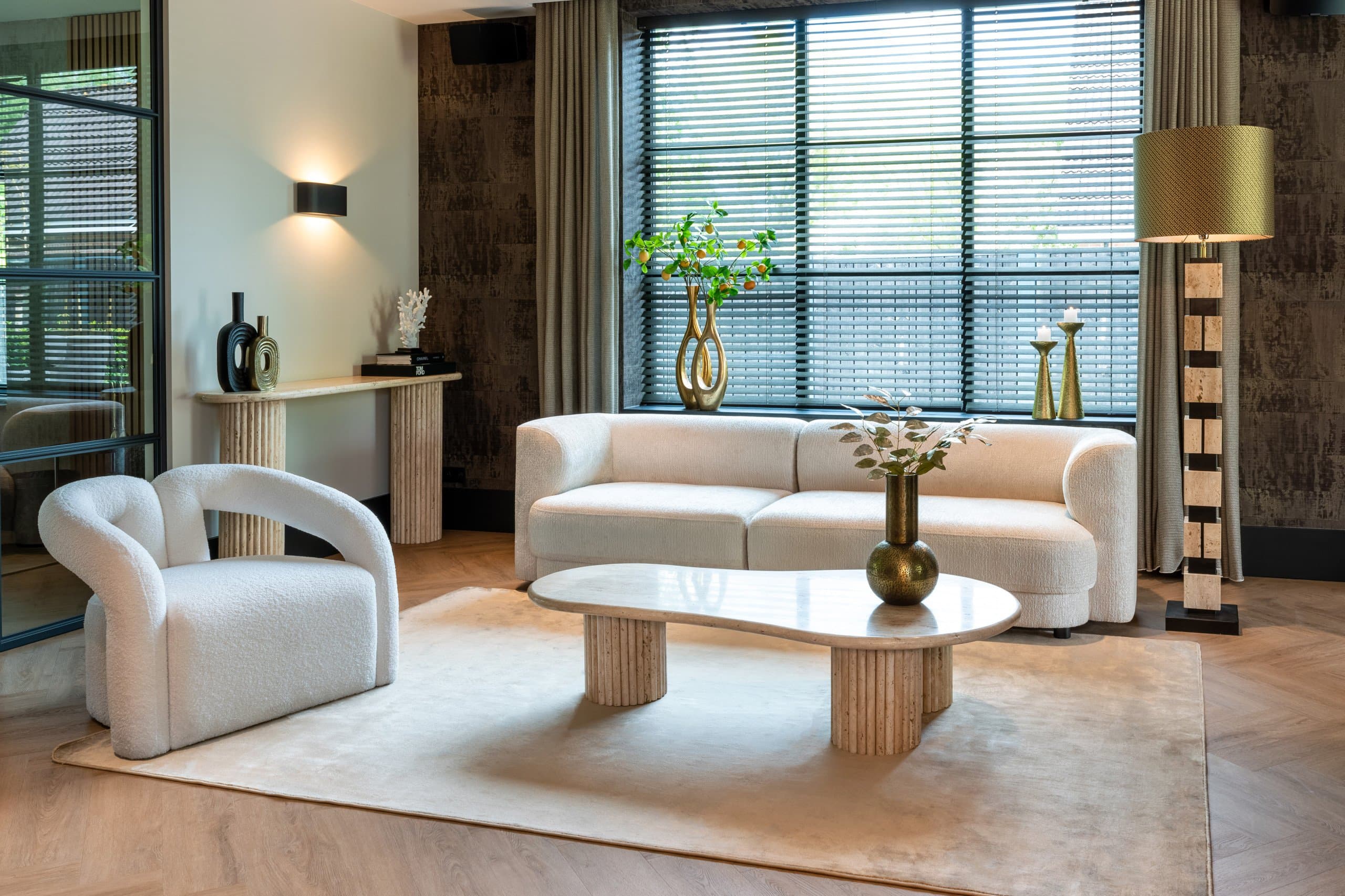 Woonkamer met een travertijnen salontafel op geribbelde zuilen, een crème bouclé bank en fauteuil, en een travertijnen console tegen de wand