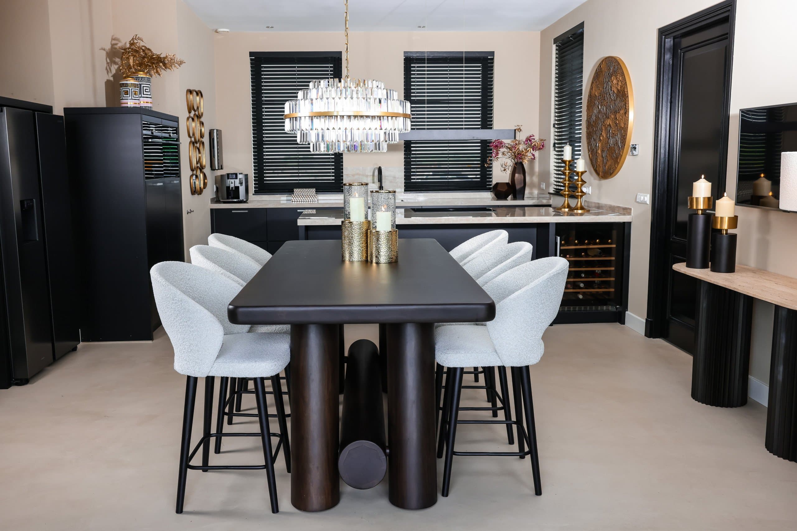 Eethoek met witte bouclé stoelen rond een donkere tafel, met op de achtergrond een keuken en twee ramen met zwarte houten jaloezieën.