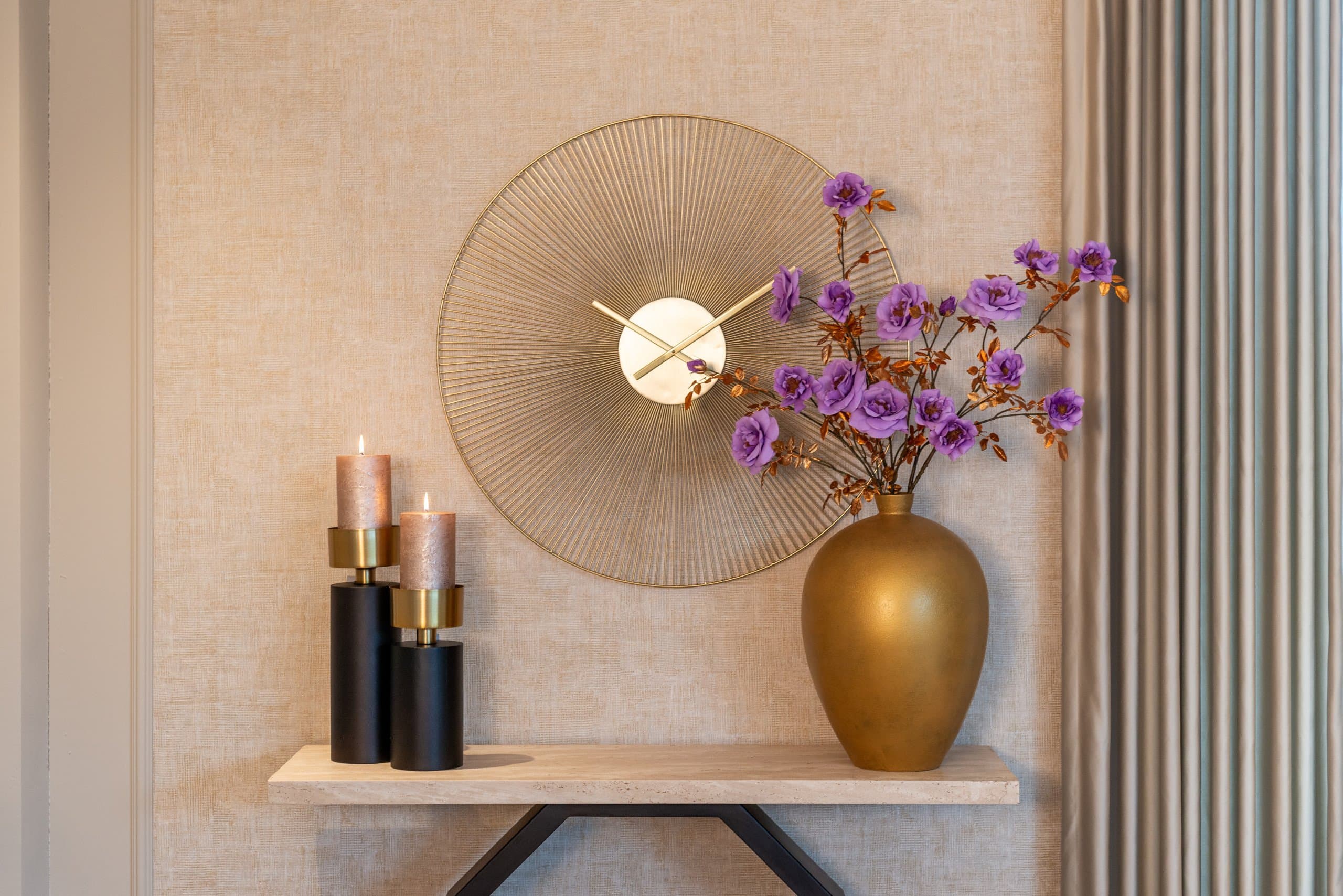 Lichte wandtafel met twee zwarte kandelaars en een gouden vaas met paarse bloemen, onder een ronde wandklok in goudkleur.