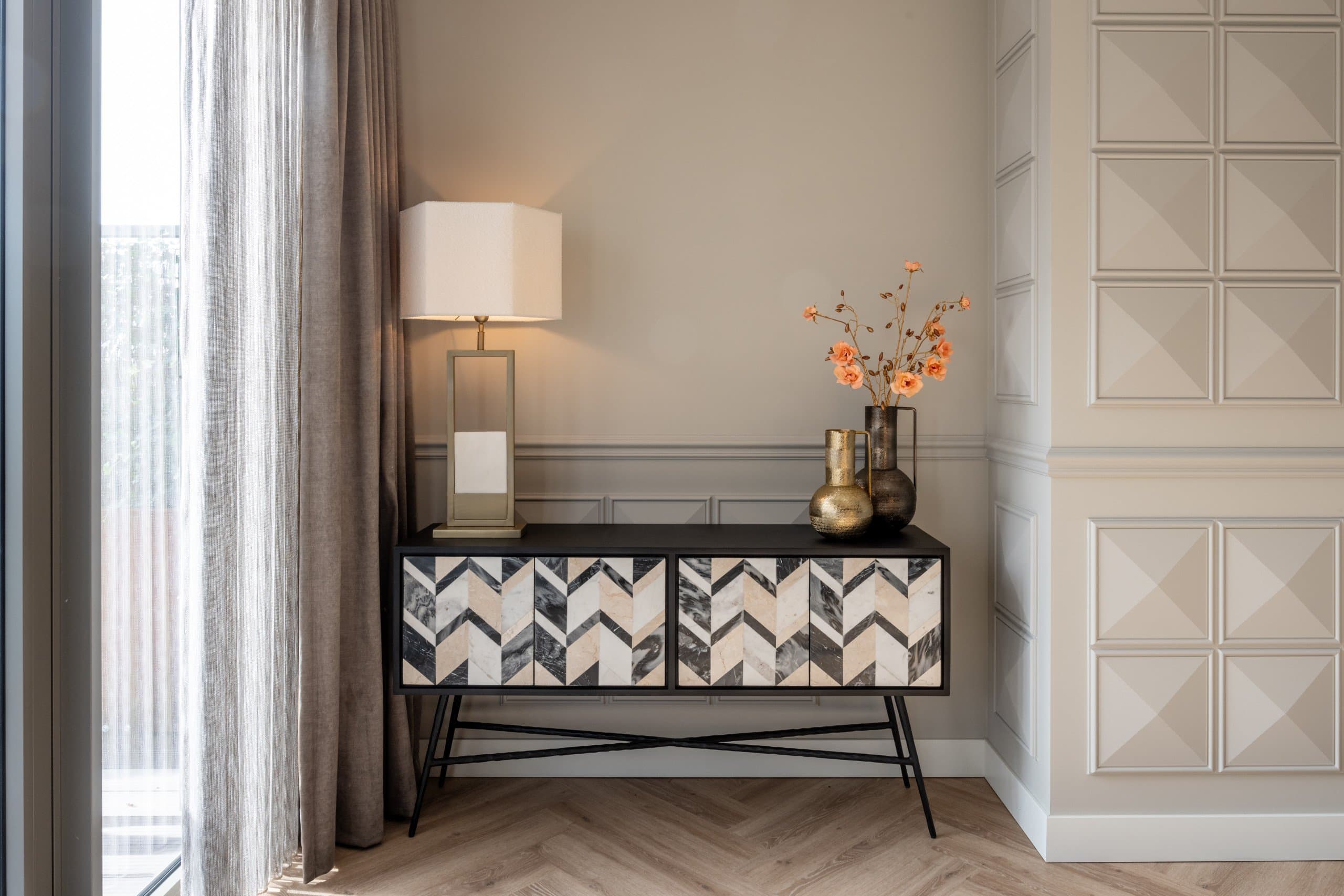 Zwarte sidetable met marmeren visgraatfronten, een tafellamp met witte kap en twee metalen vazen met oranje bloemen tegen een beige lambriseringswand