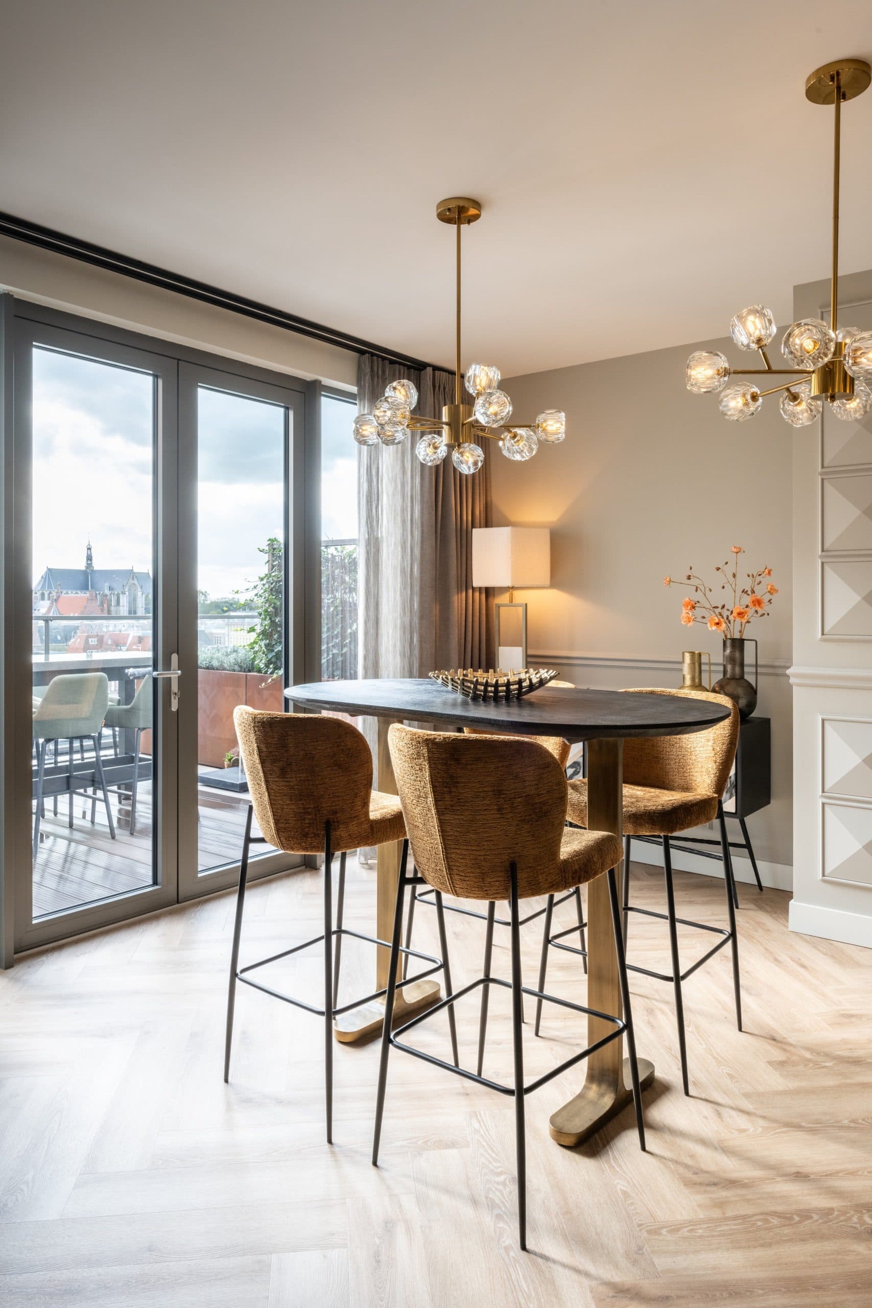 Ovale donkere bartafel op een goudkleurig metalen onderstel met vier okerkleurige barstoelen, voor openslaande deuren met uitzicht op een balkon