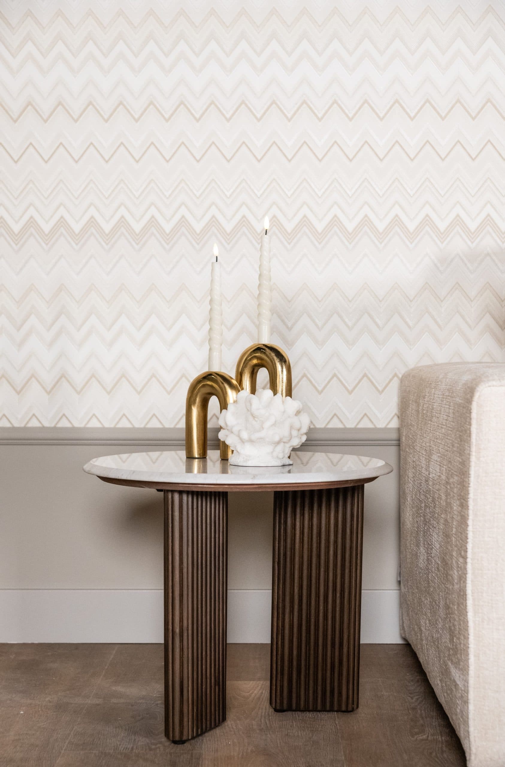 Ronde bijzettafel met wit marmeren blad en geribbelde houten voet naast een lichte bank, met twee gouden kandelaars en brandende witte kaarsen erop