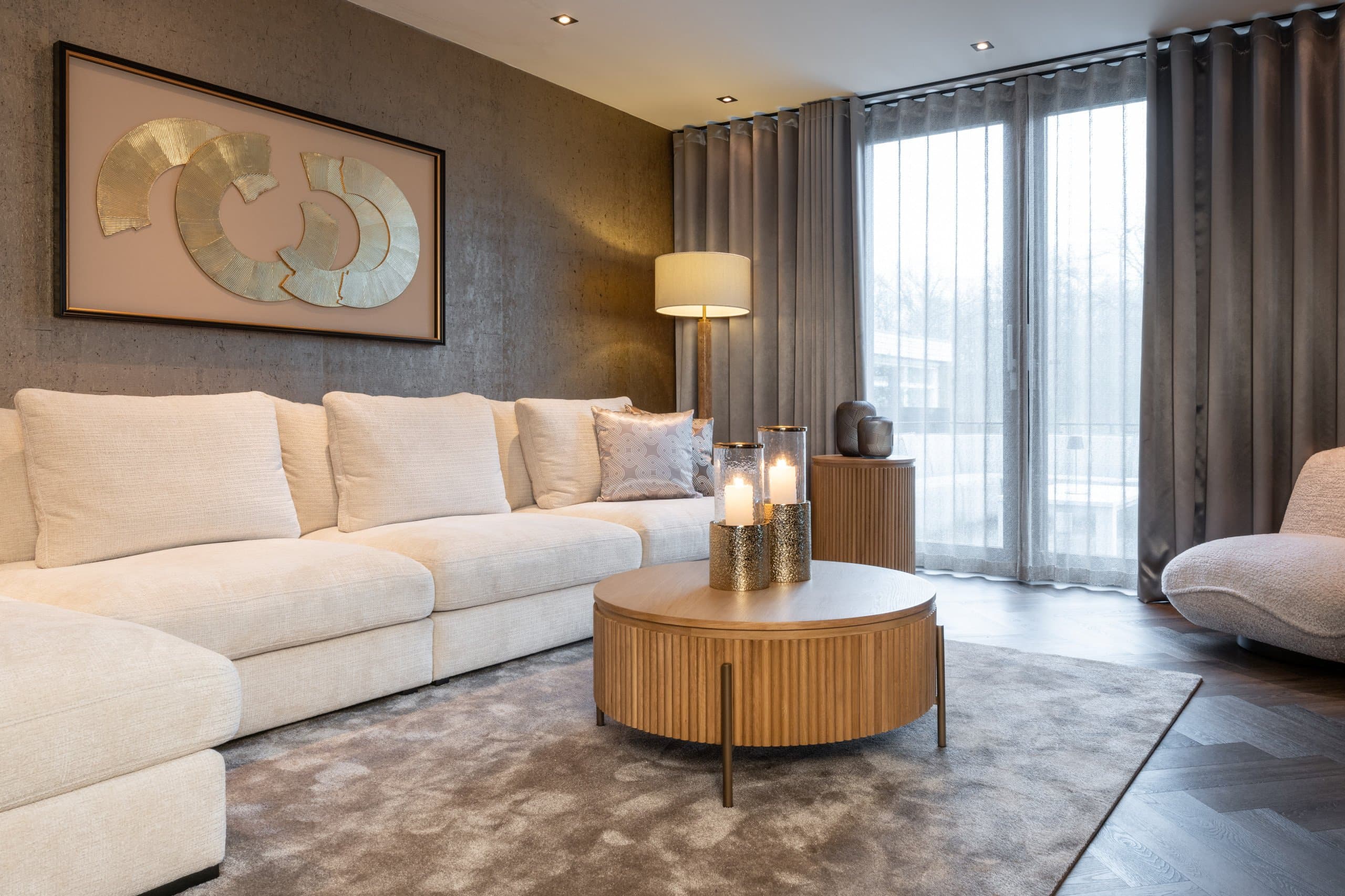 Avondlijke woonkamer met een crèmekleurige bank, een brandende staande lamp met crème kap en drie aangestoken kaarsen in glazen windlichten op een ronde houten salontafel