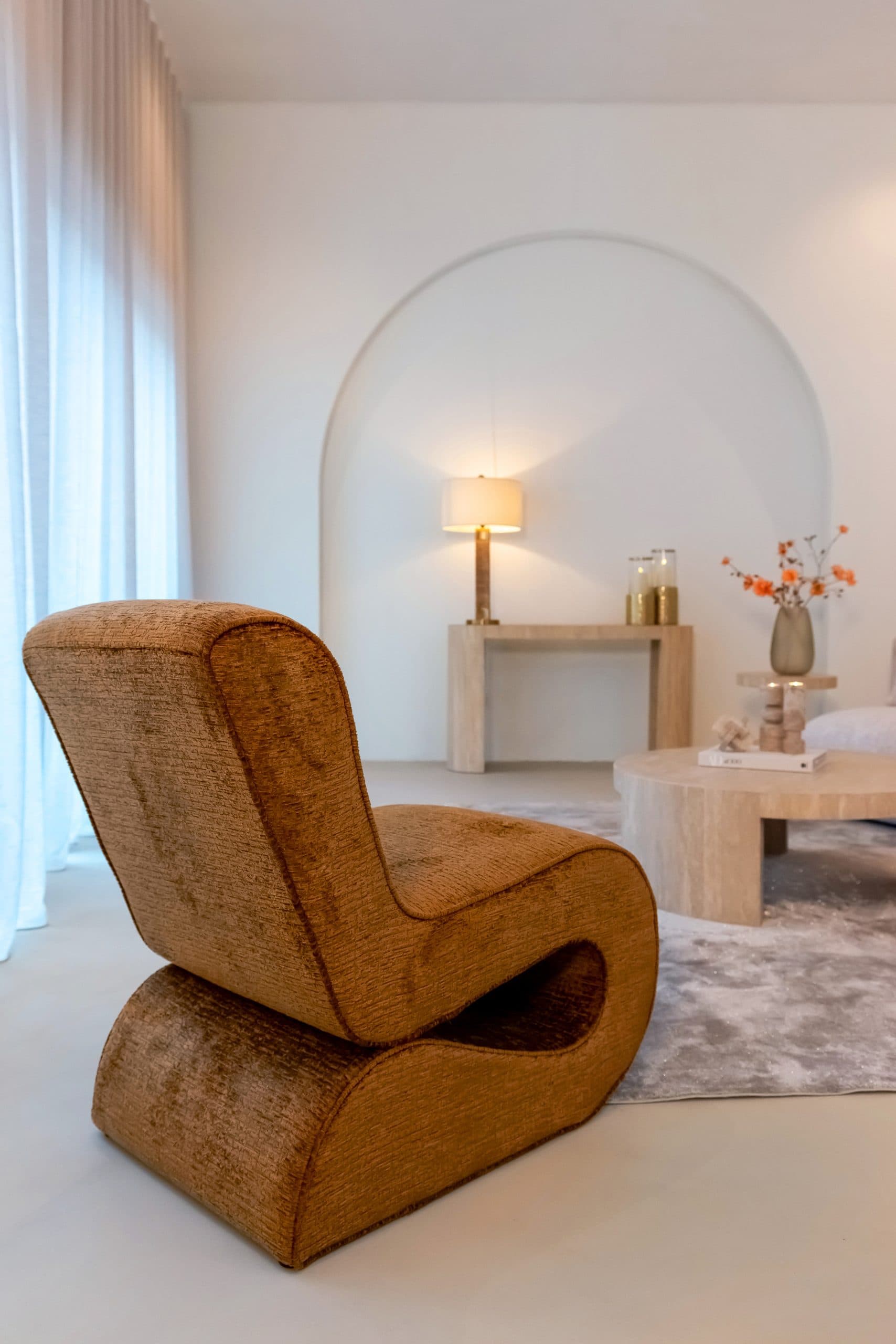 Cognackleurige fauteuil met hoge gebogen rug op de voorgrond, met daarachter een travertijnen console met tafellamp en windlichten