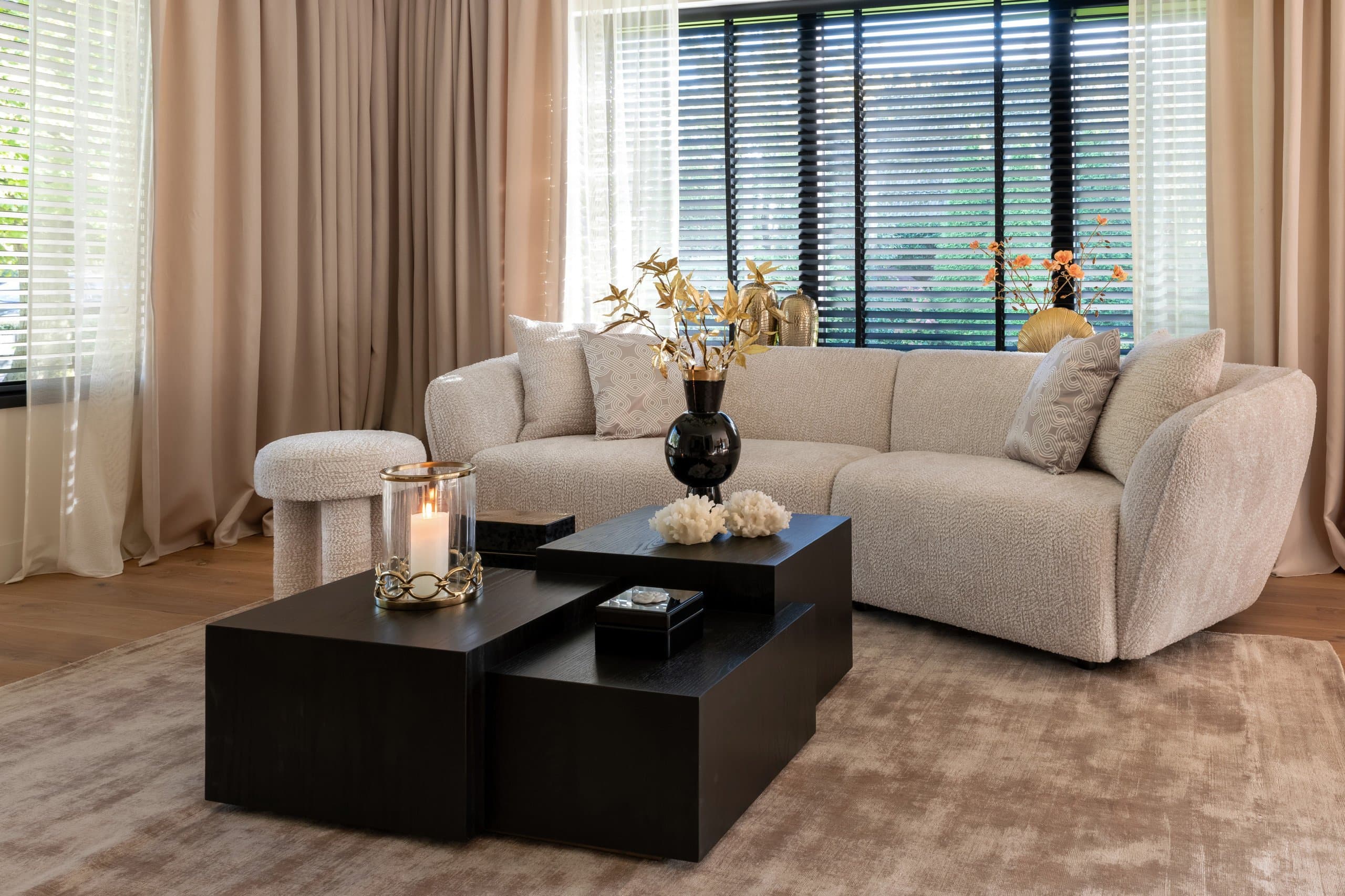 Crèmekleurige bouclé bank met sierkussens in een lichte woonkamer, met zwarte salontafels, een windlicht en beige gordijnen voor de ramen
