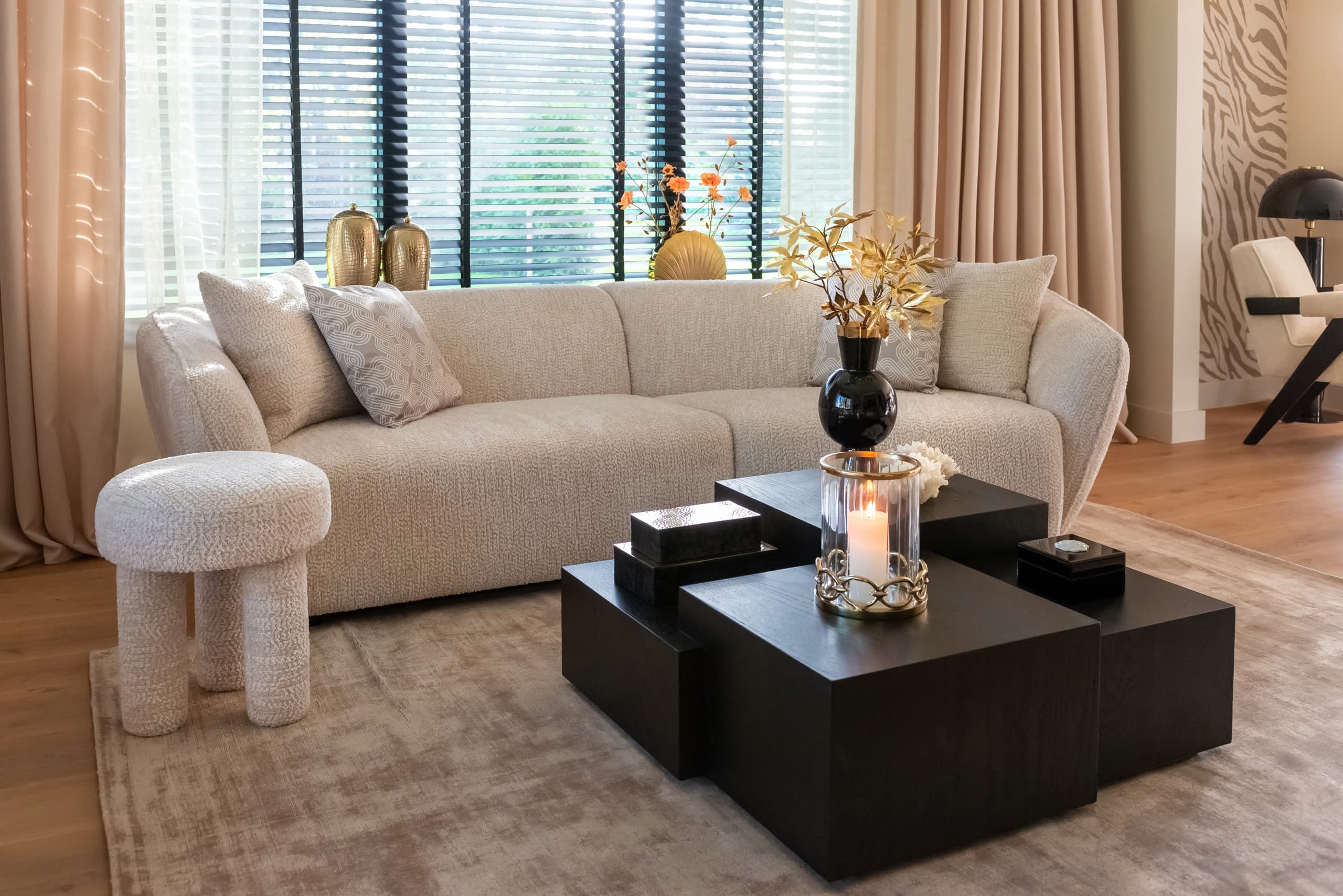Woonkamer met een crèmekleurige bouclébank, een ronde poef, zwarte vierkante salontafels met een kaars en een vaas, op een beige vloerkleed voor een raam met jaloezieën.