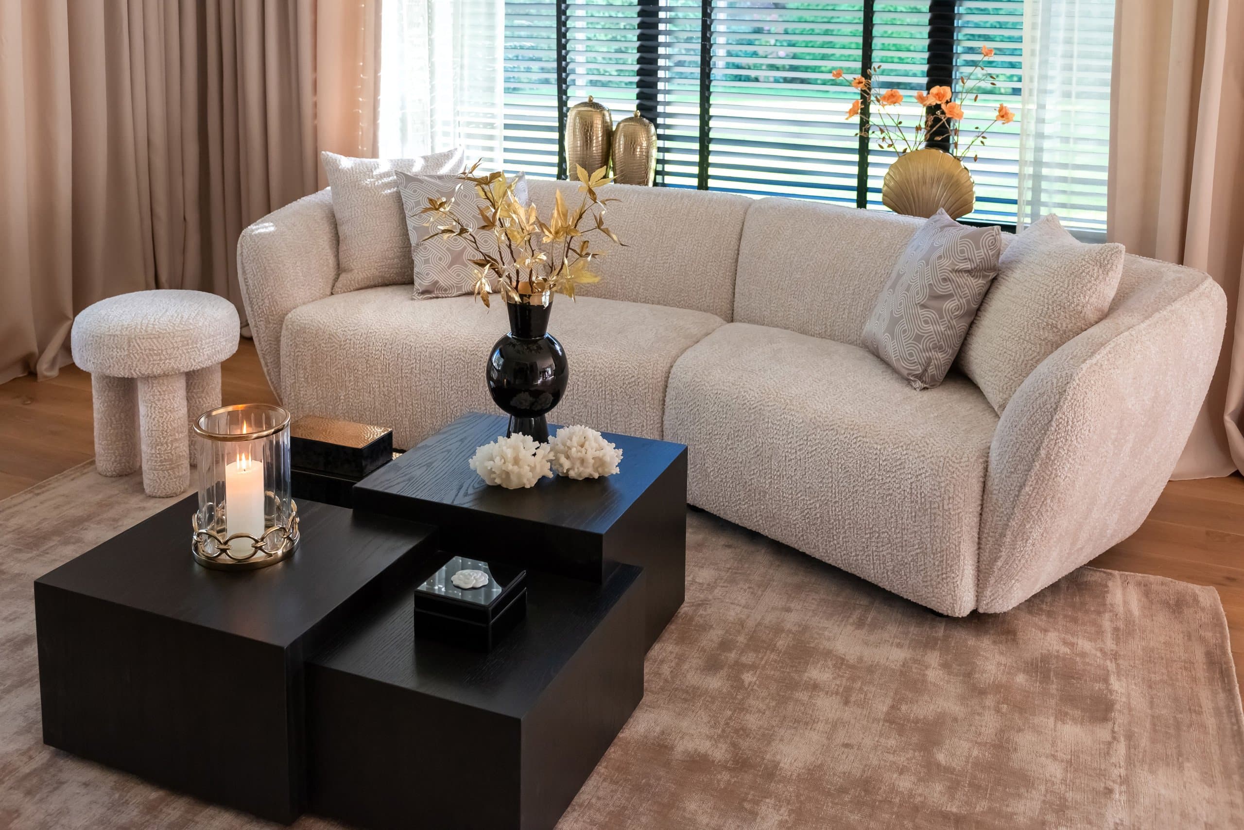 Woonkamer met een lage crèmekleurige bouclébank, twee zwarte salontafels, een beige vloerkleed en gordijnen die van het plafond tot de vloer vallen