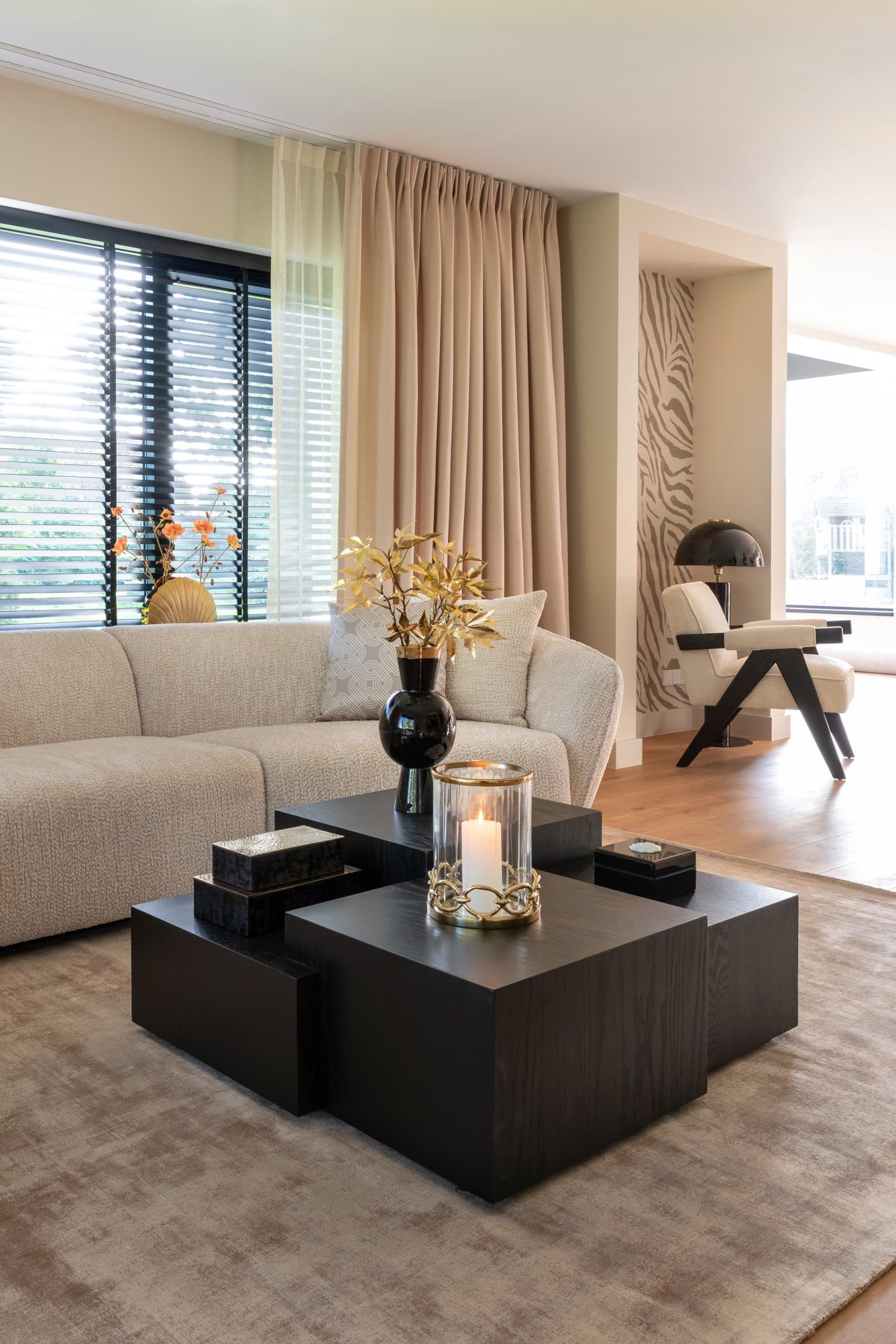 Woonkamer met een crèmekleurige bouclébank, zwarte salontafels met een brandende kaars in een windlicht en op de achtergrond een fauteuil met een zwarte tafellamp bij het raam.