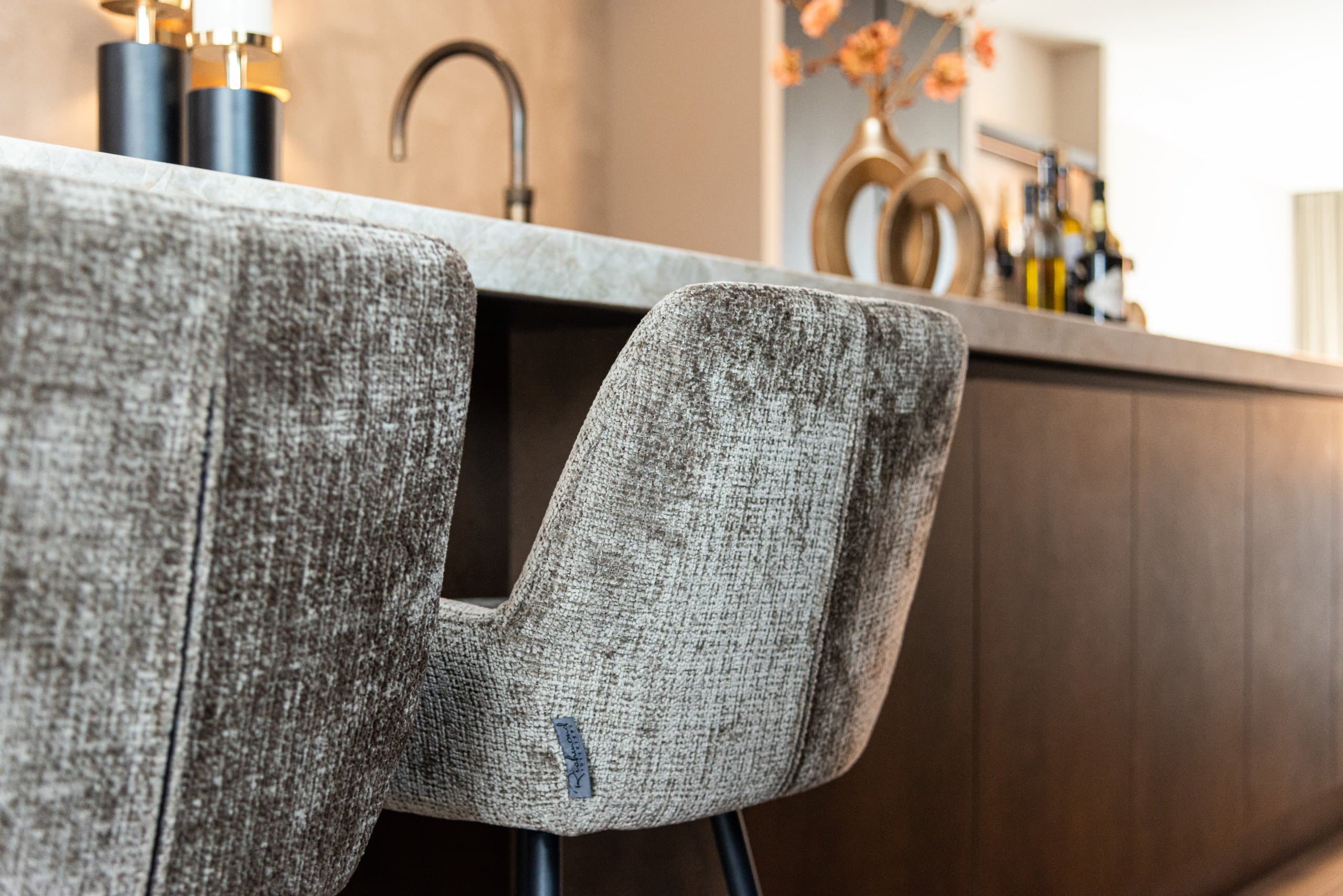 Twee gestoffeerde counterstoelen in grijze chenille aan een keukeneiland met een stenen blad, met op de achtergrond een kraan en donkere keukenkasten