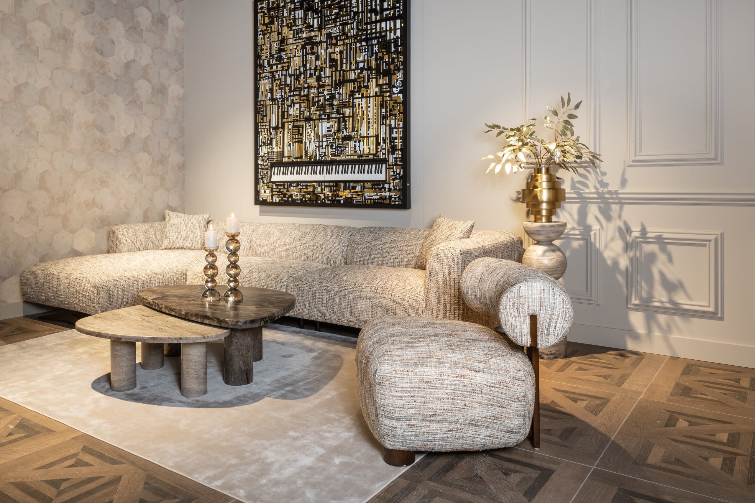 Gouden vaas met zijden bloemen en groene takken op een stenen sokkel tegen een witte paneelwand, met een beige bank en travertin bijzettafels ervoor.