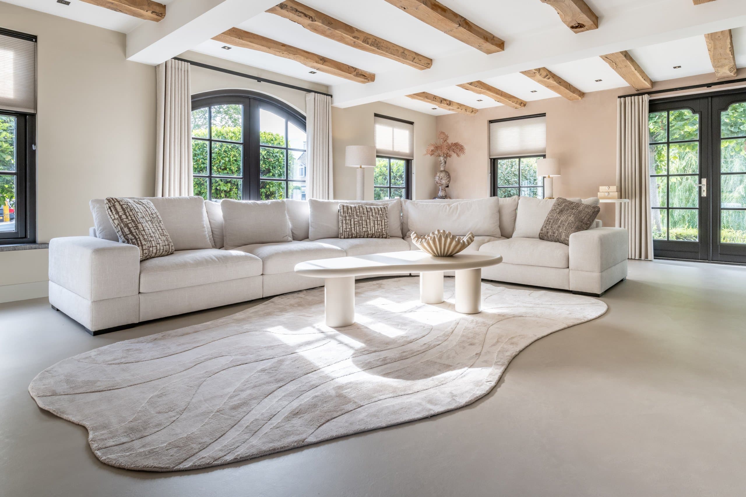 Groot beige vloerkleed met golvende rand in een lichte woonkamer met witte hoekbank en ronde salontafel