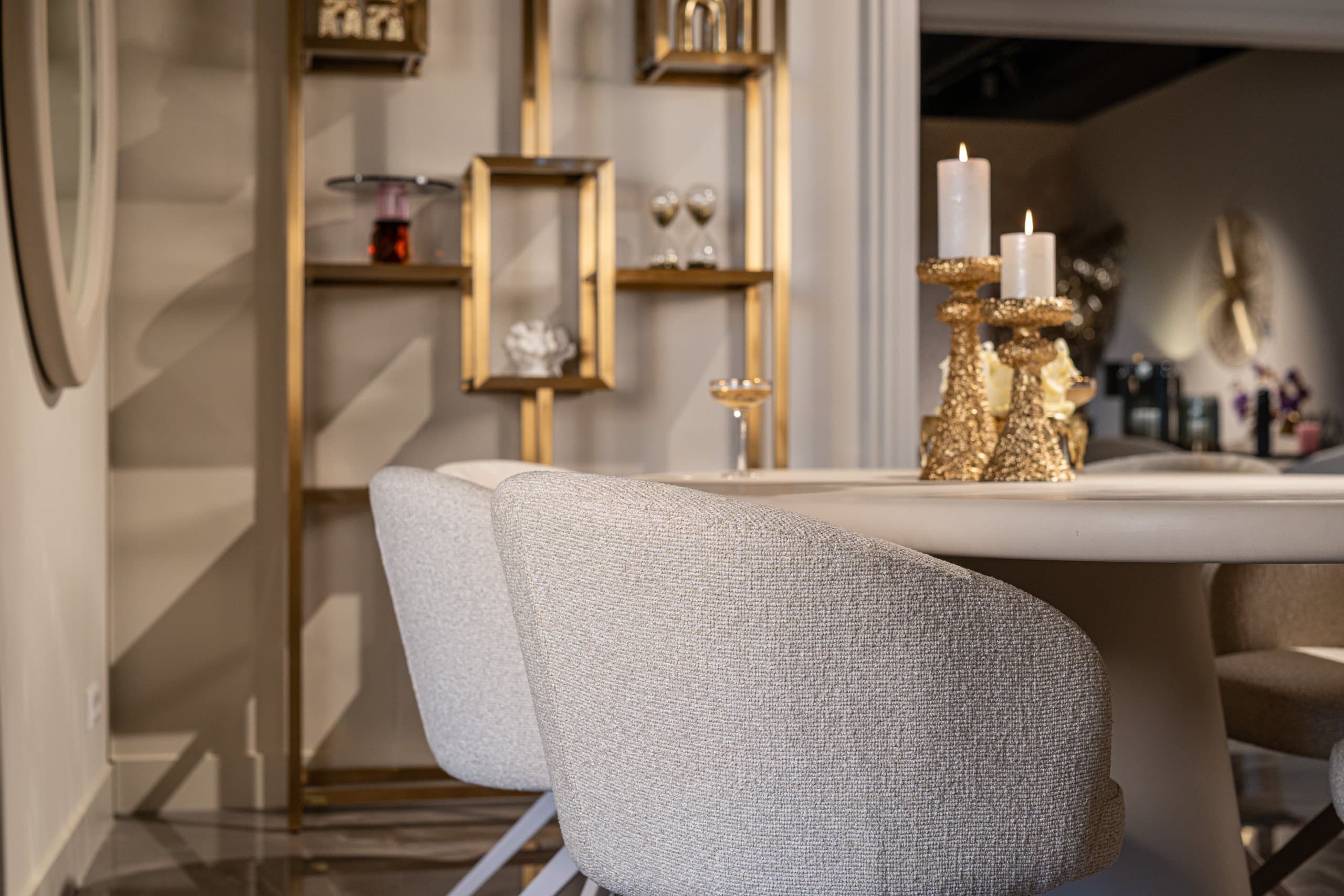 Eethoek met lichte gestoffeerde stoelen aan een ronde tafel met twee gouden kandelaars, en op de achtergrond een open goudkleurige vitrinekast
