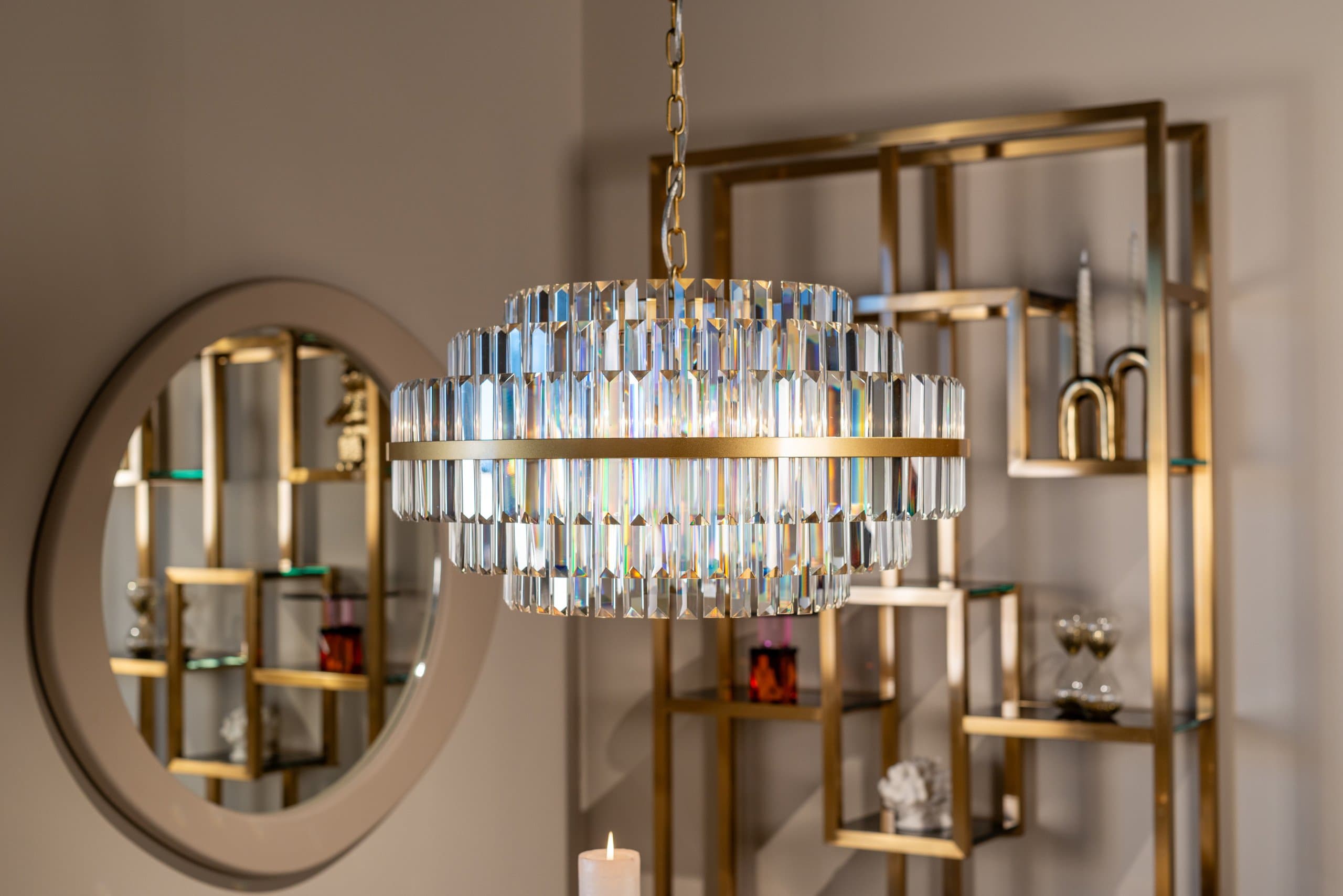 Ronde wandspiegel met een brede matte beige lijst naast een kristallen hanglamp, met op de achtergrond een goudkleurige open vakkenkast
